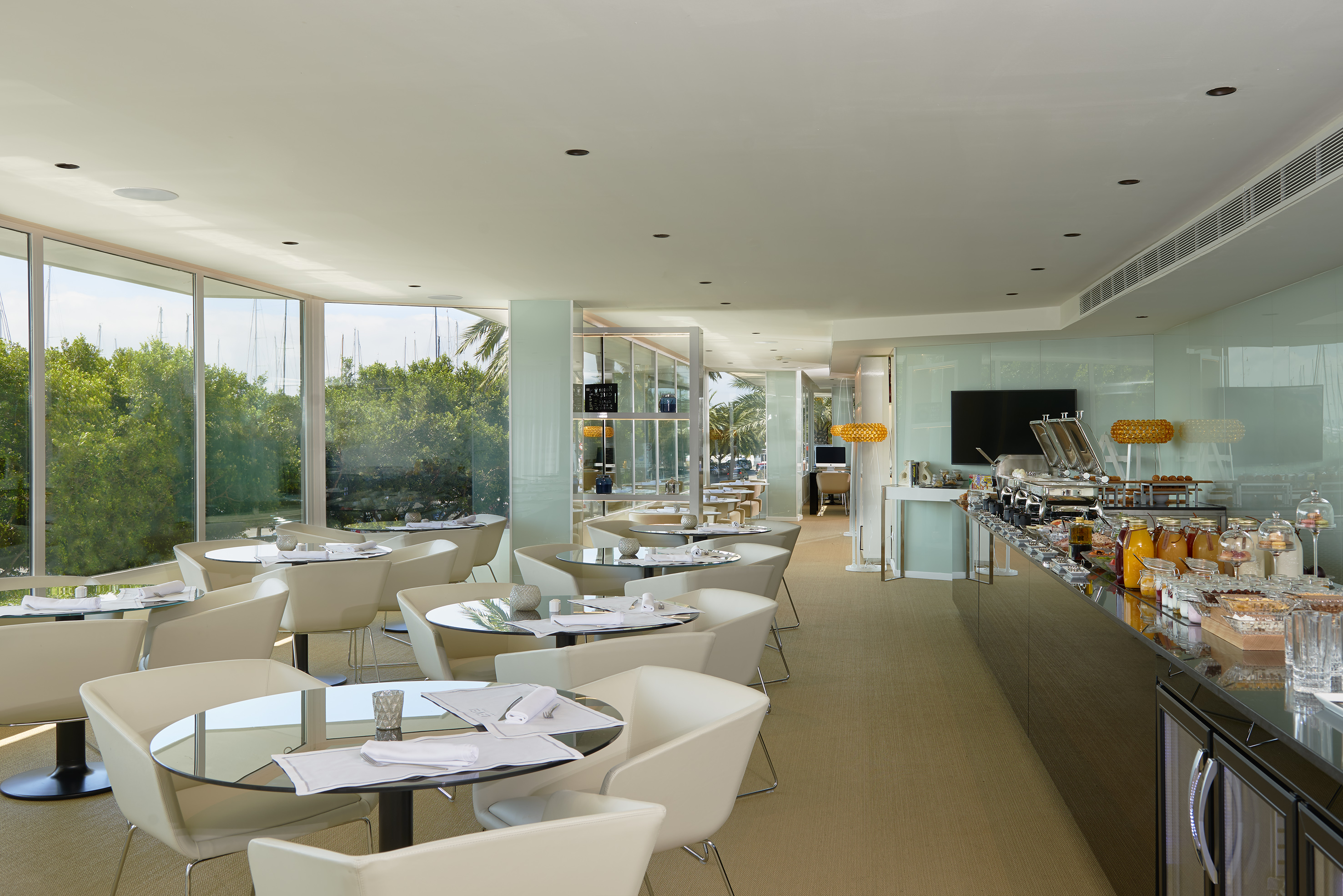 Sleek hotel restaurant with breakfast buffet, cream chairs, and marina view.