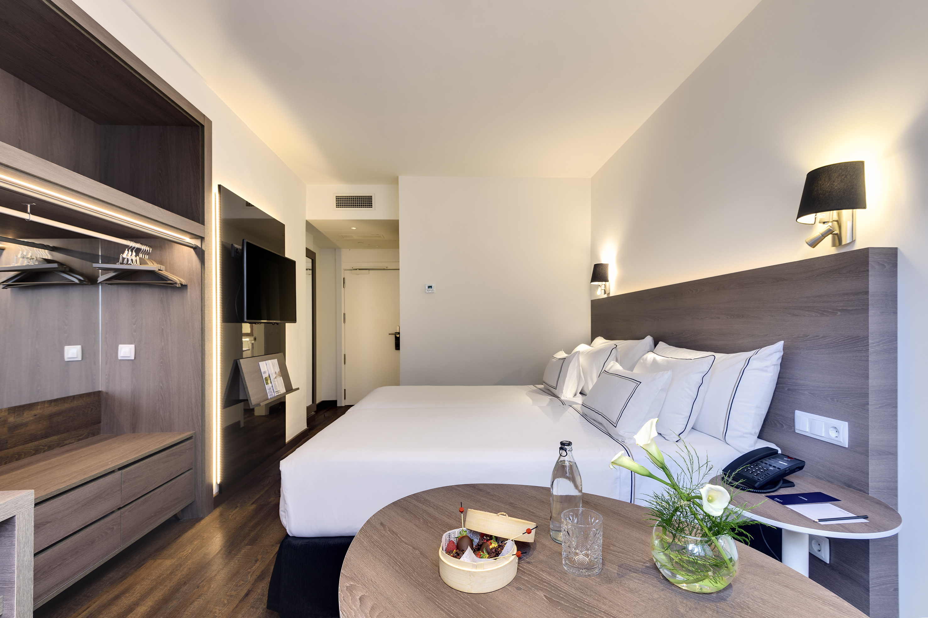 Modern hotel room with large bed, wooden wardrobe, and round bedside table.