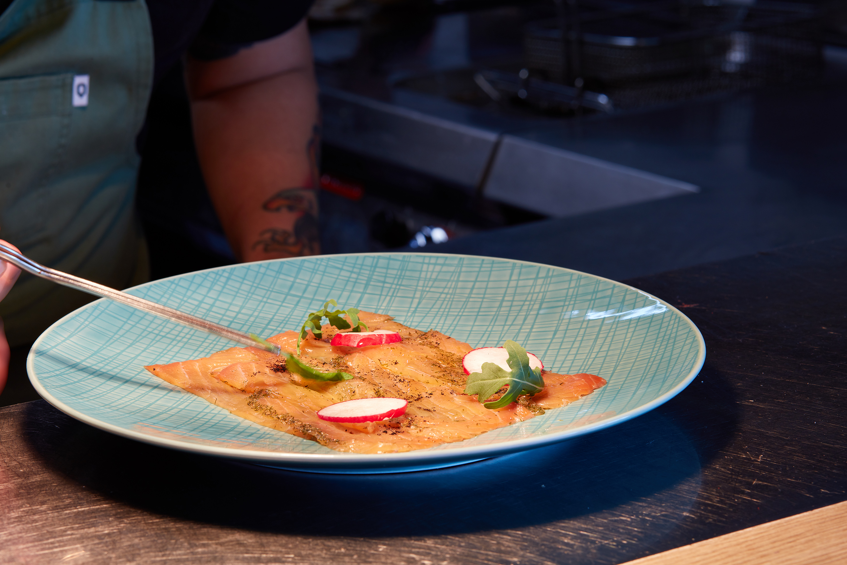 Chef plates smoked salmon, radishes, and herbs on a patterned blue plate.