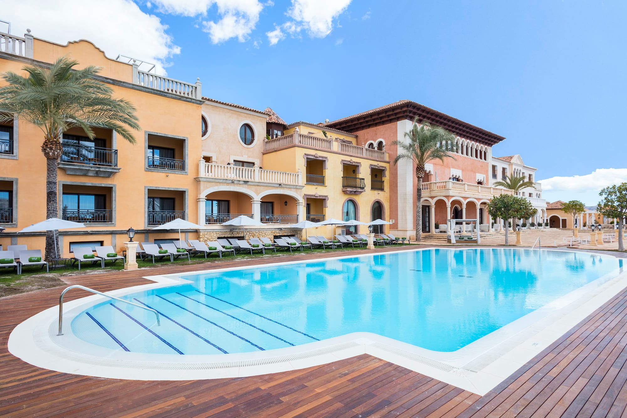 Mediterranean resort with large outdoor pool, wooden deck, and palm trees.