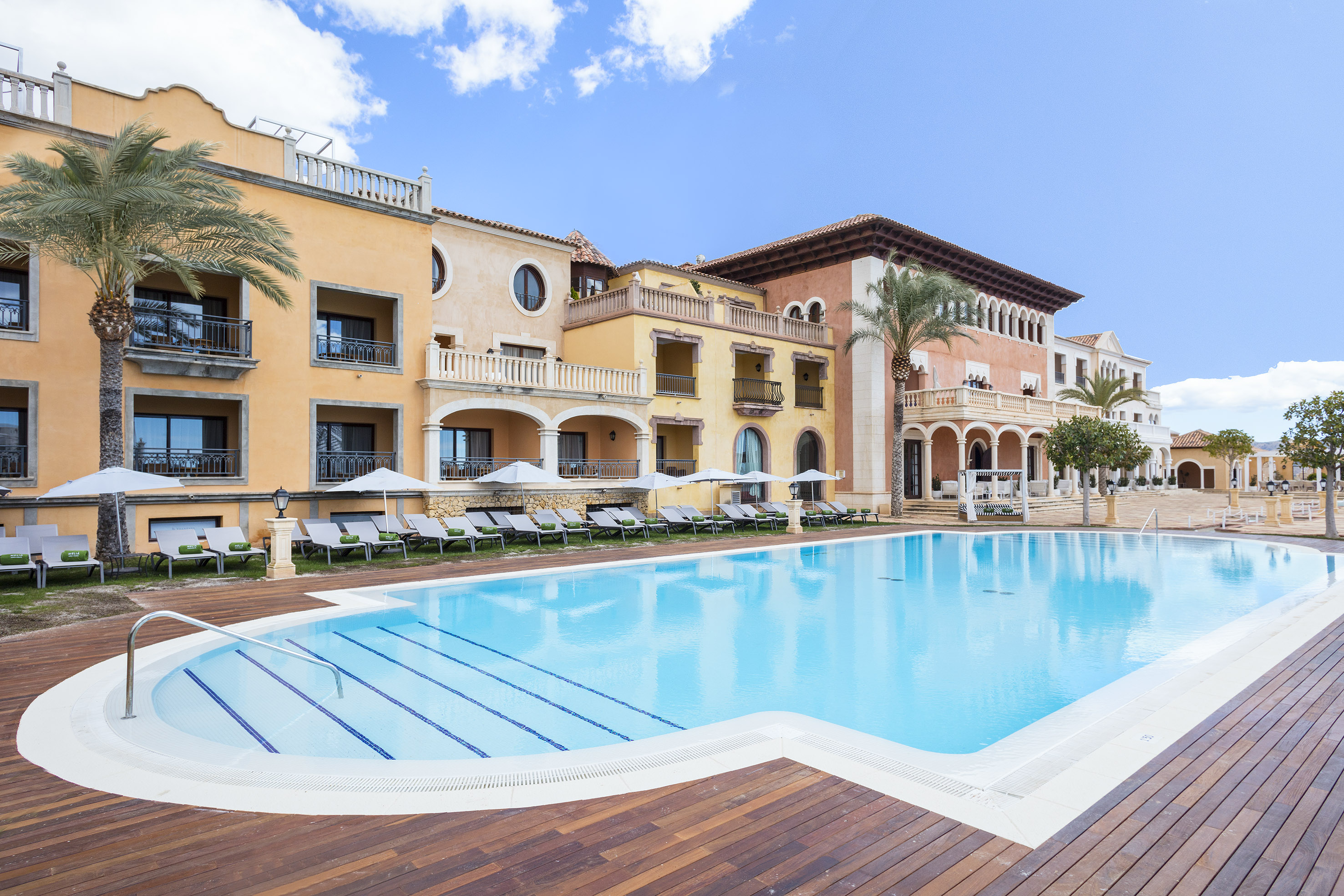 Mediterranean resort with large outdoor pool, wooden deck, and palm trees.