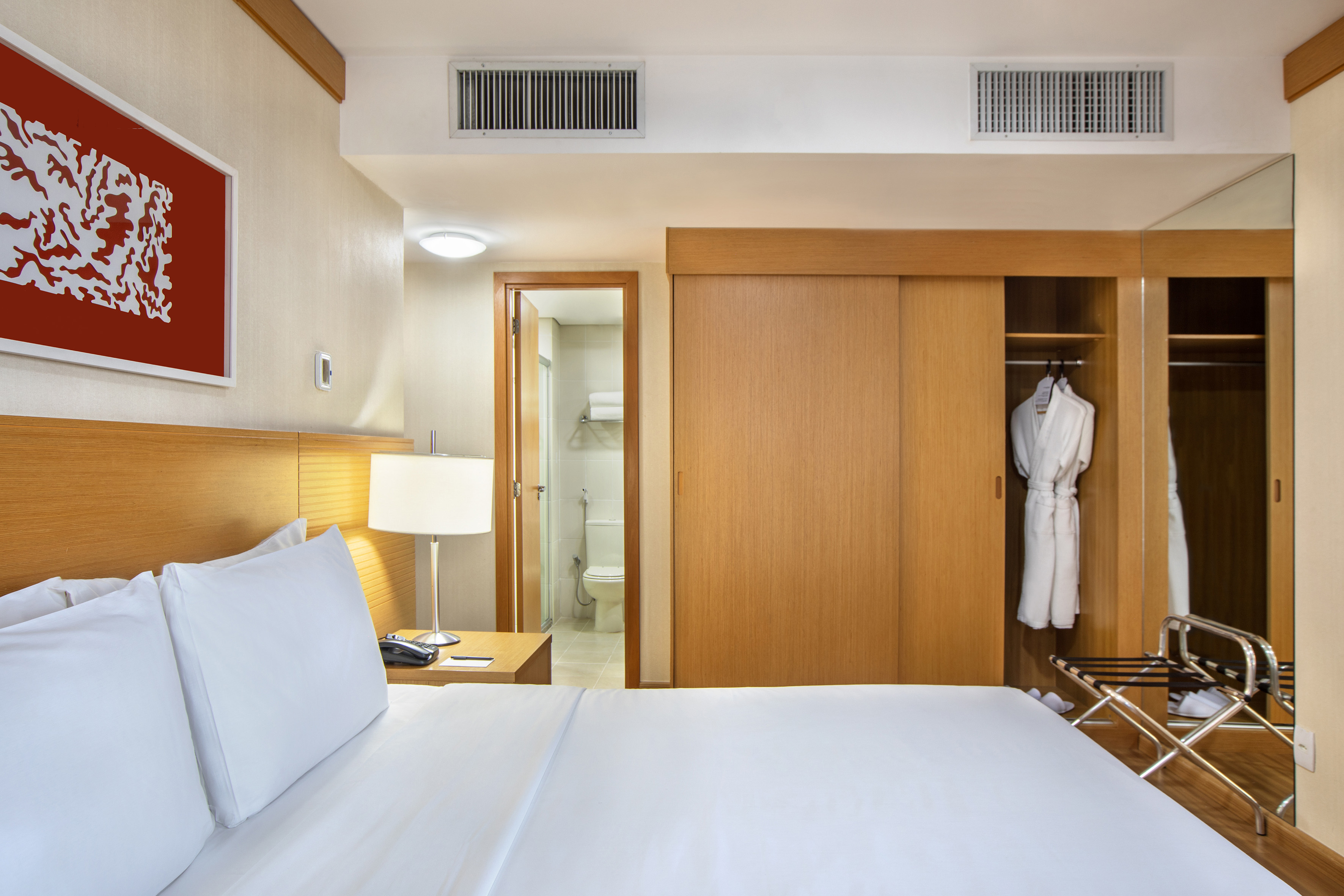 Clean hotel room with white bed, wooden closet, red wall art, and bathroom view.