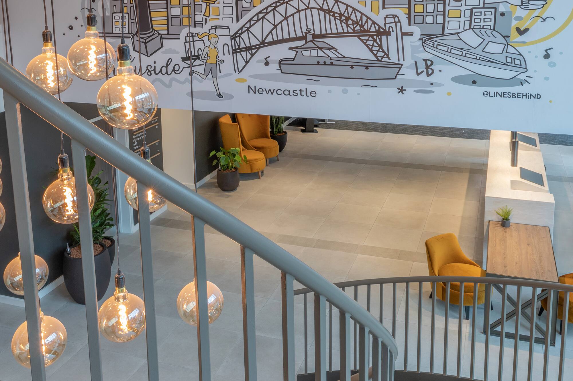 Hotel lobby featuring Newcastle mural, spiral staircase, and hanging Edison lights.