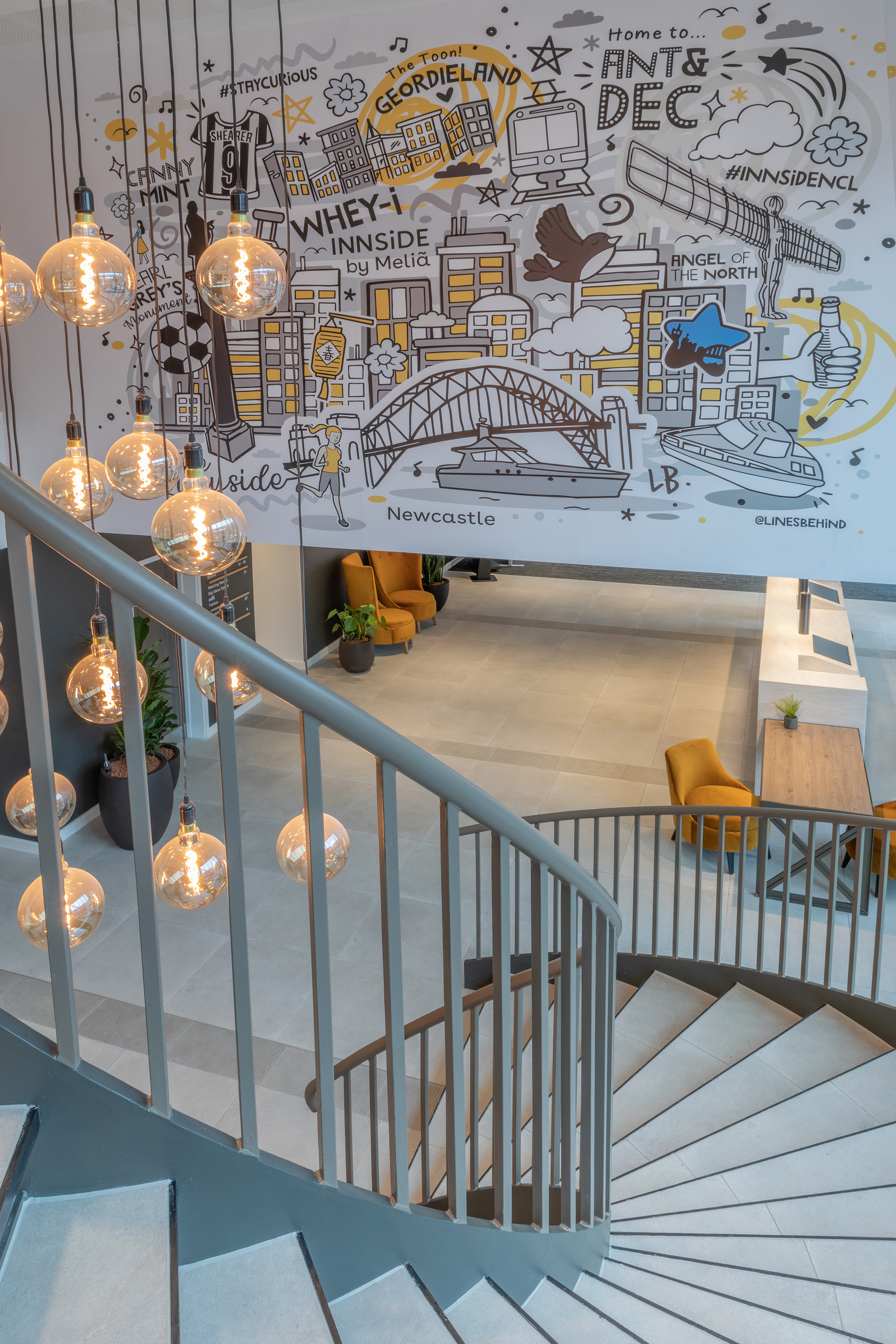 Hotel lobby featuring Newcastle mural, spiral staircase, and hanging Edison lights.