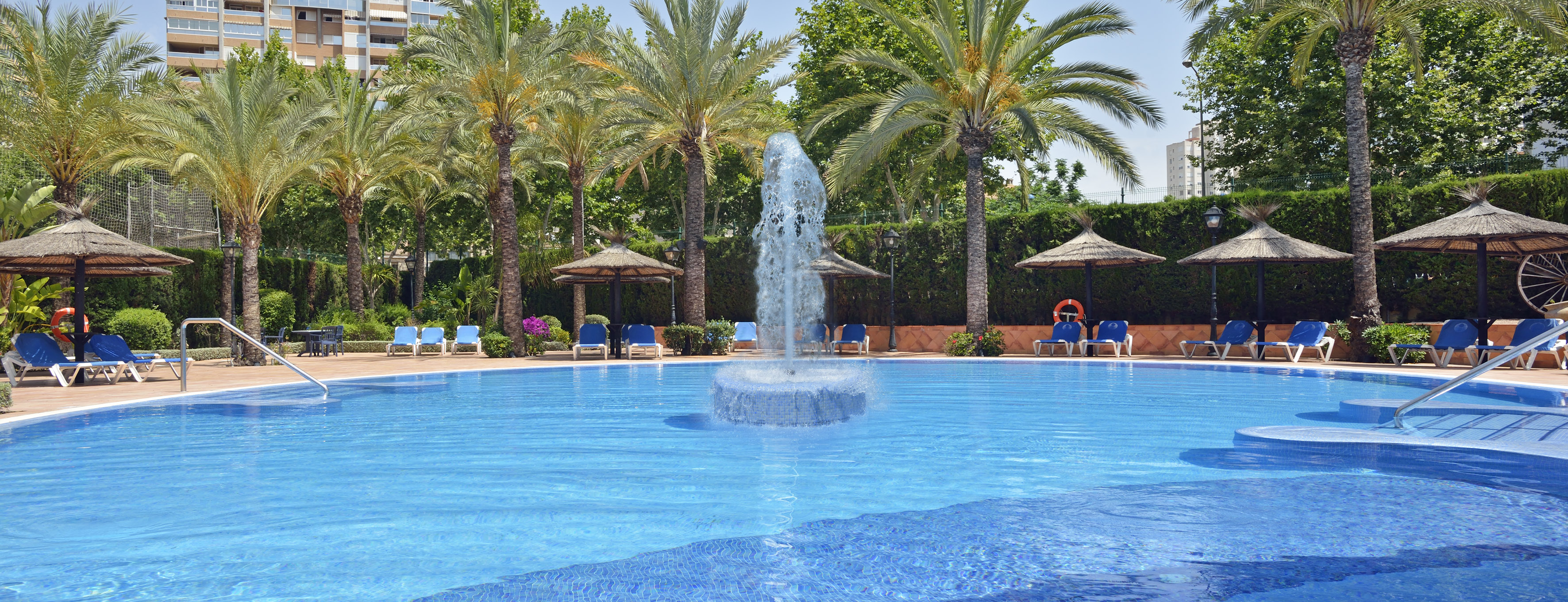 Sunny resort pool with fountain, palm trees, and blue lounge chairs.