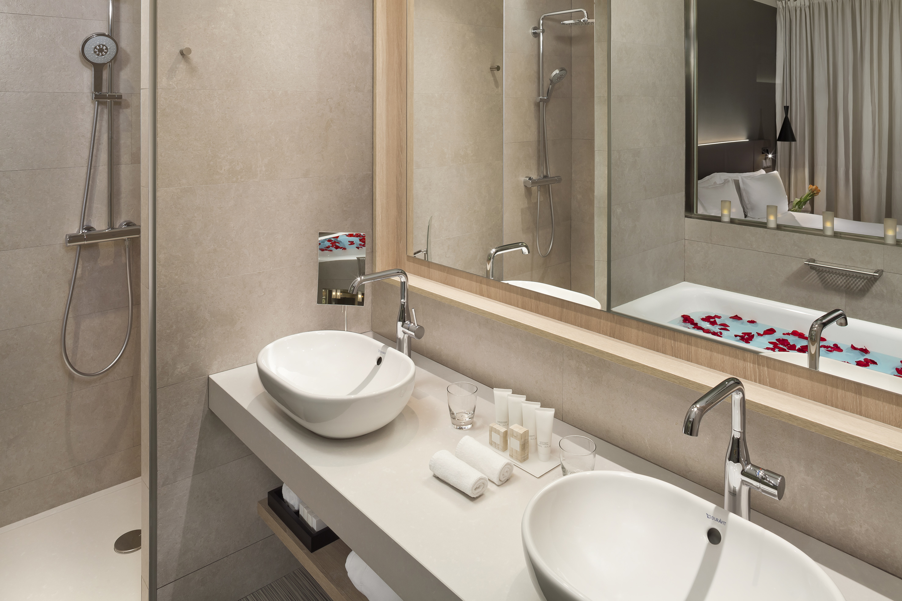 Modern hotel bathroom with dual sinks, shower, and reflected tub with rose petals.