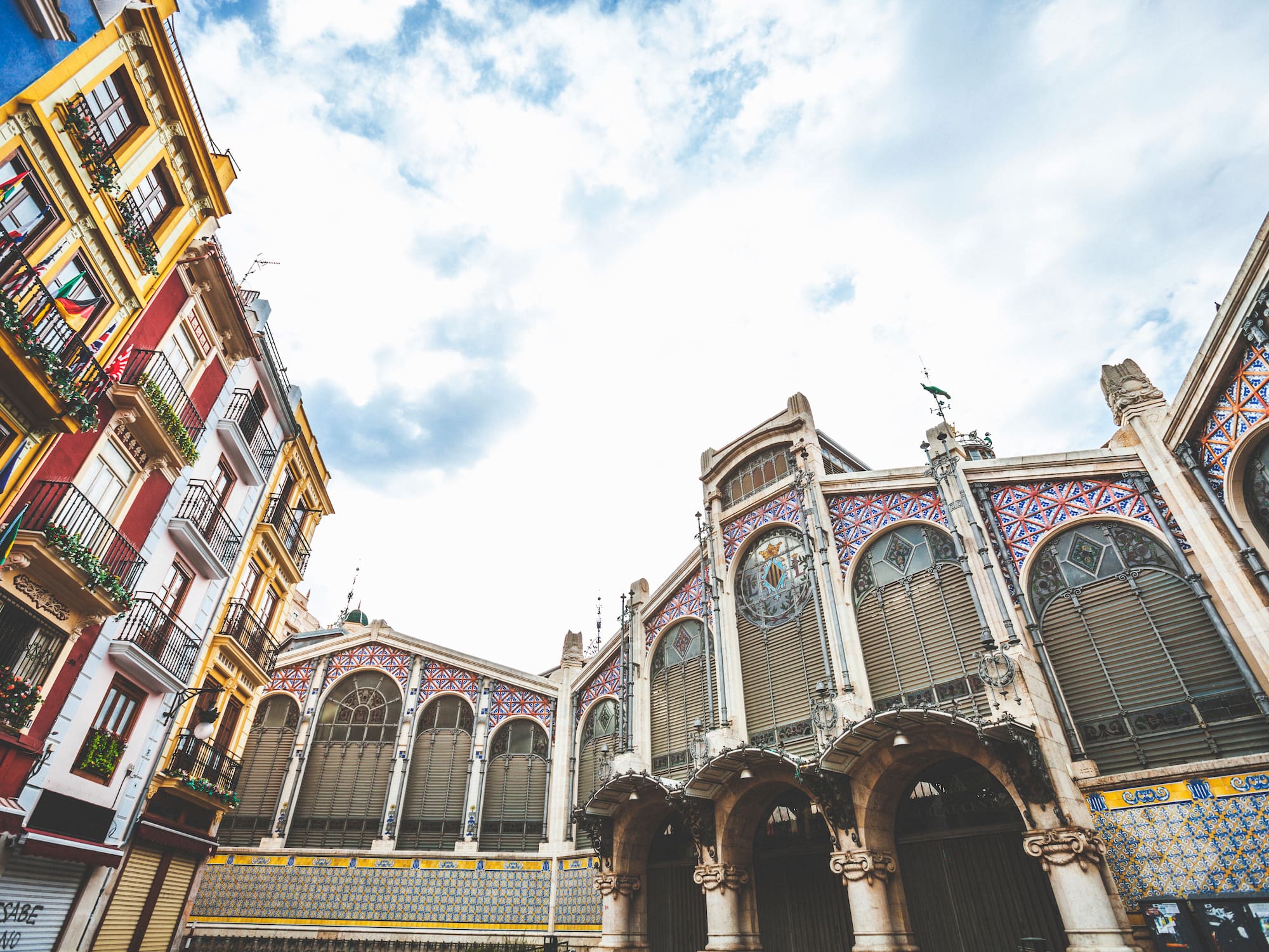 Ornate Valencia Central Market architecture next to colorful city buildings under cloudy sky.
