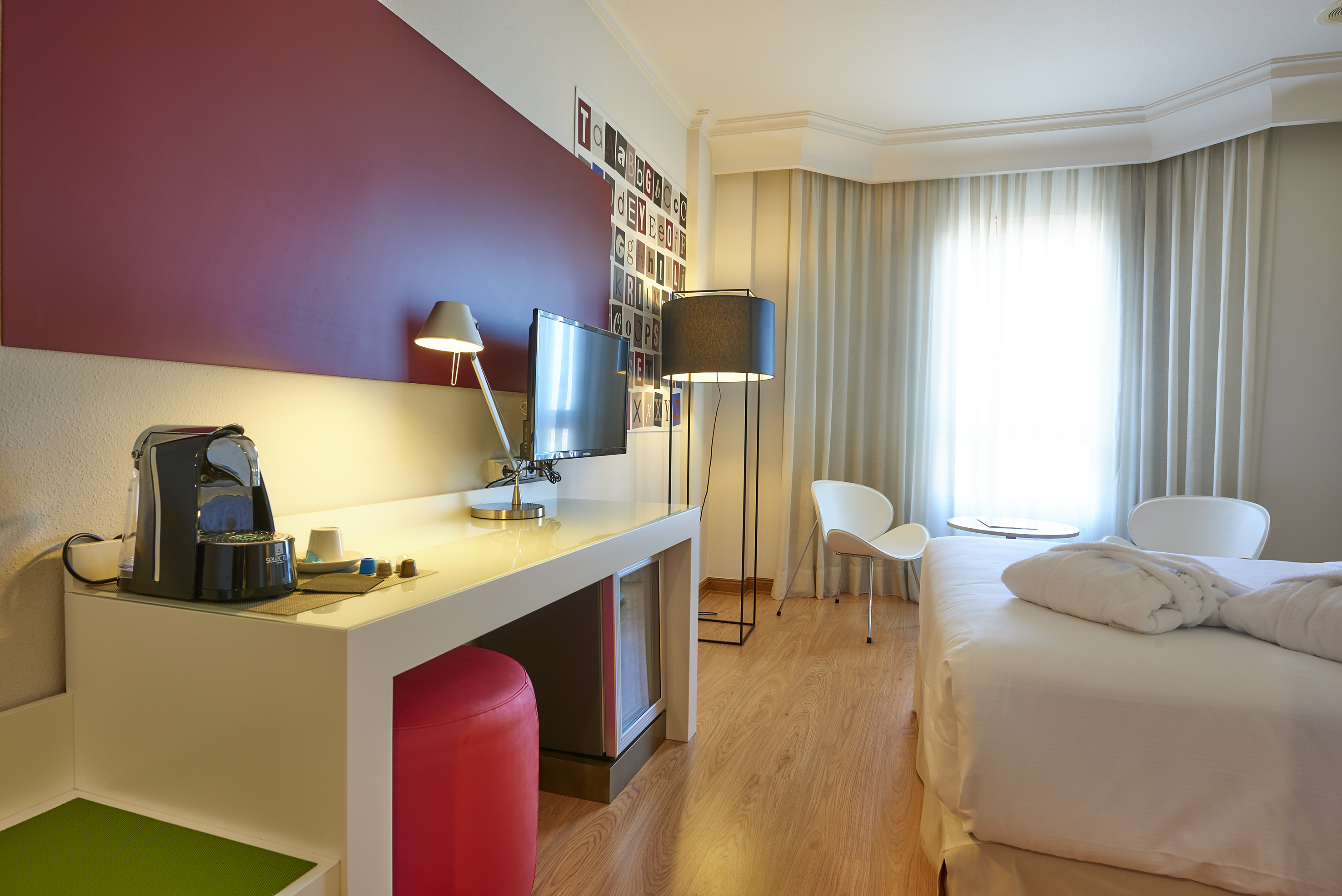 Modern hotel room with coffee machine, desk, TV, bed, and bright window.