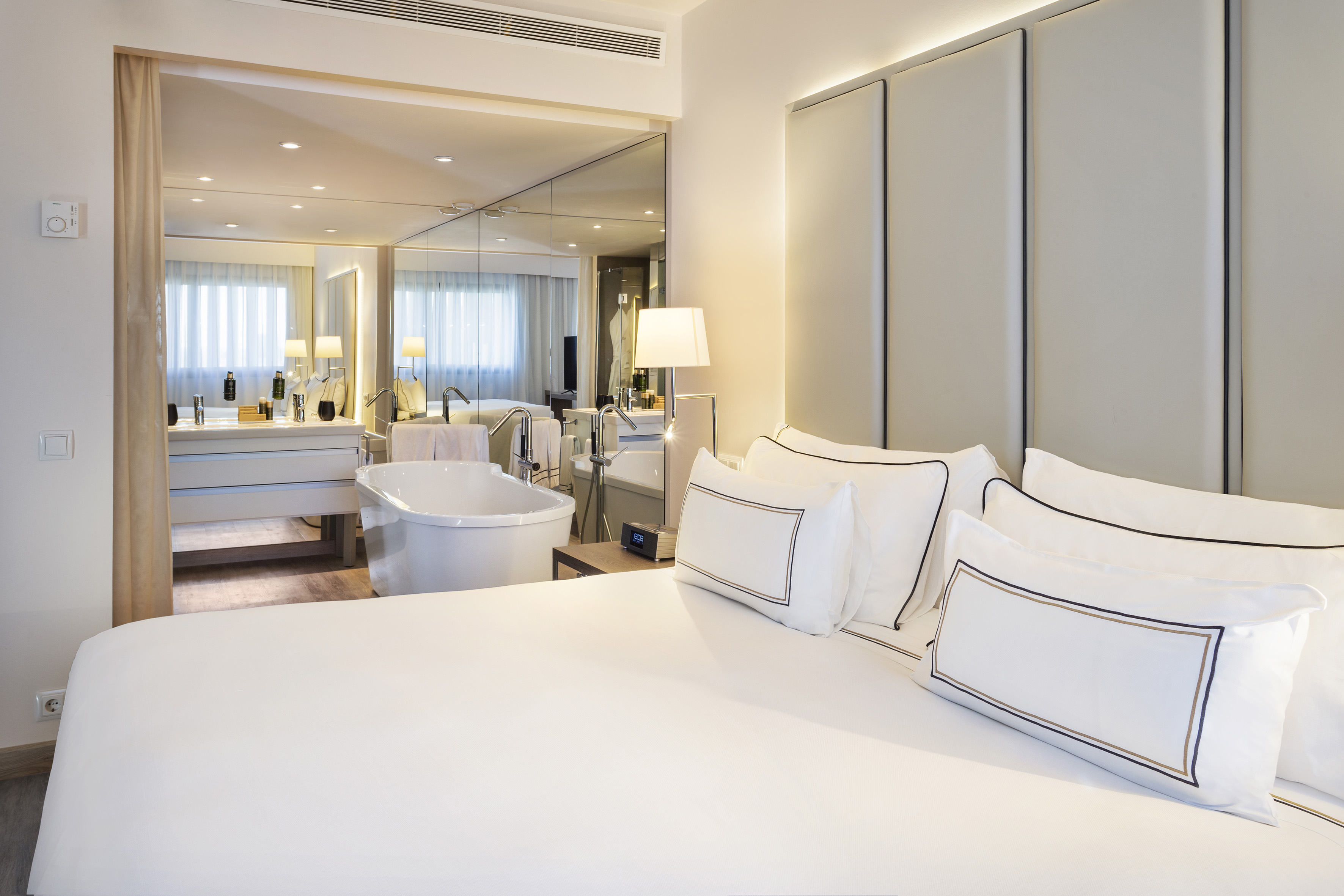 Elegant hotel suite with king bed, mirrored wall, and freestanding bathtub.