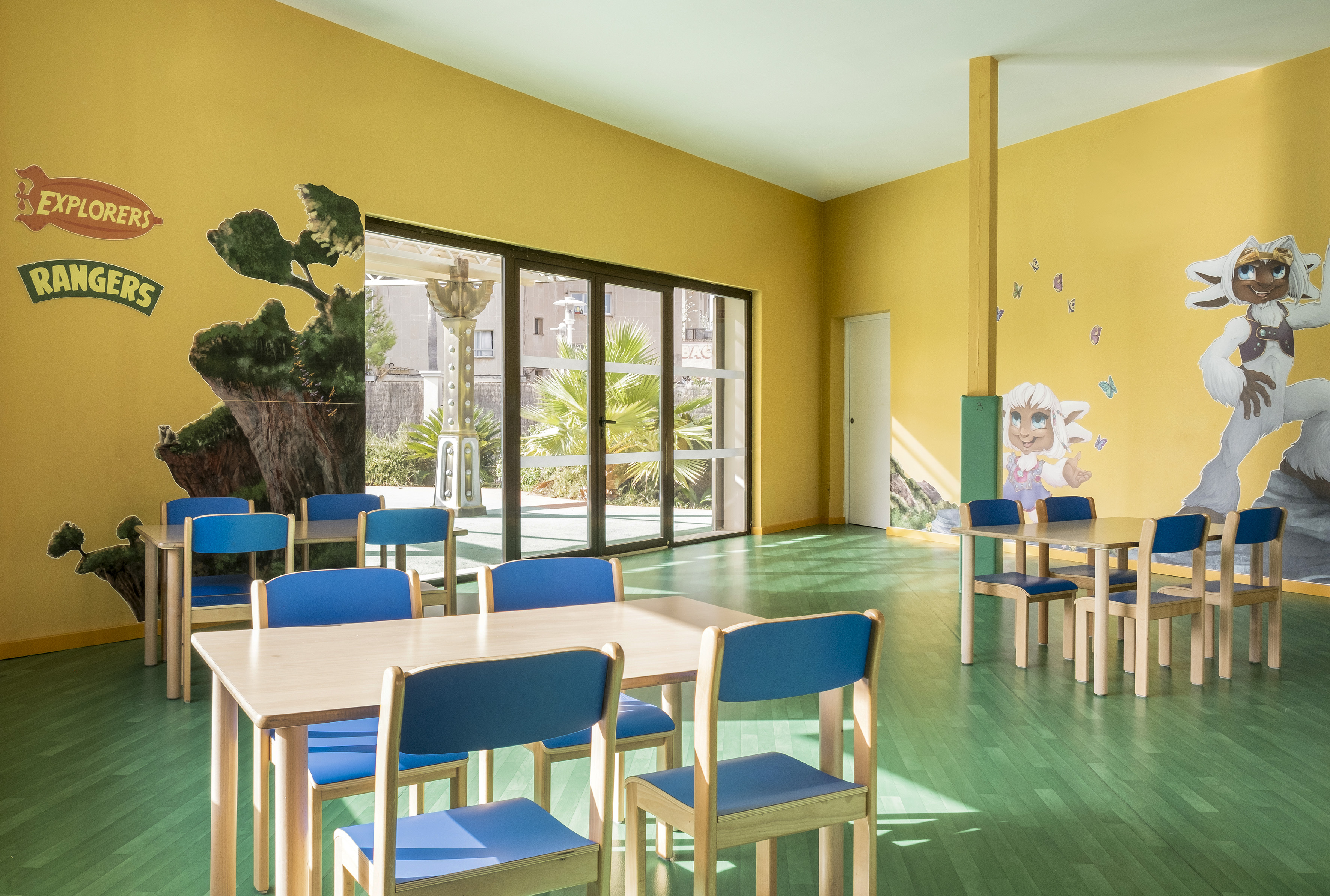Bright yellow kids' club room with green floor, murals, wooden tables, and blue chairs.