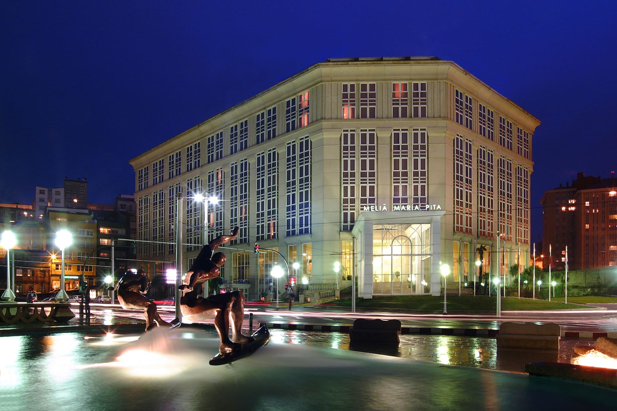 Melia Maria Pita hotel illuminated at night, fountain sculptures in city square.
