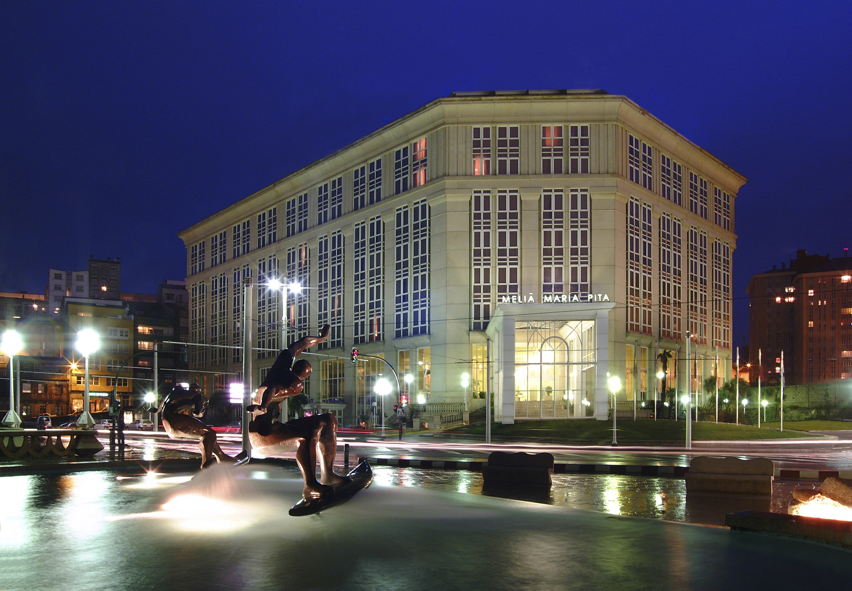 Melia Maria Pita hotel illuminated at night, fountain sculptures in city square.