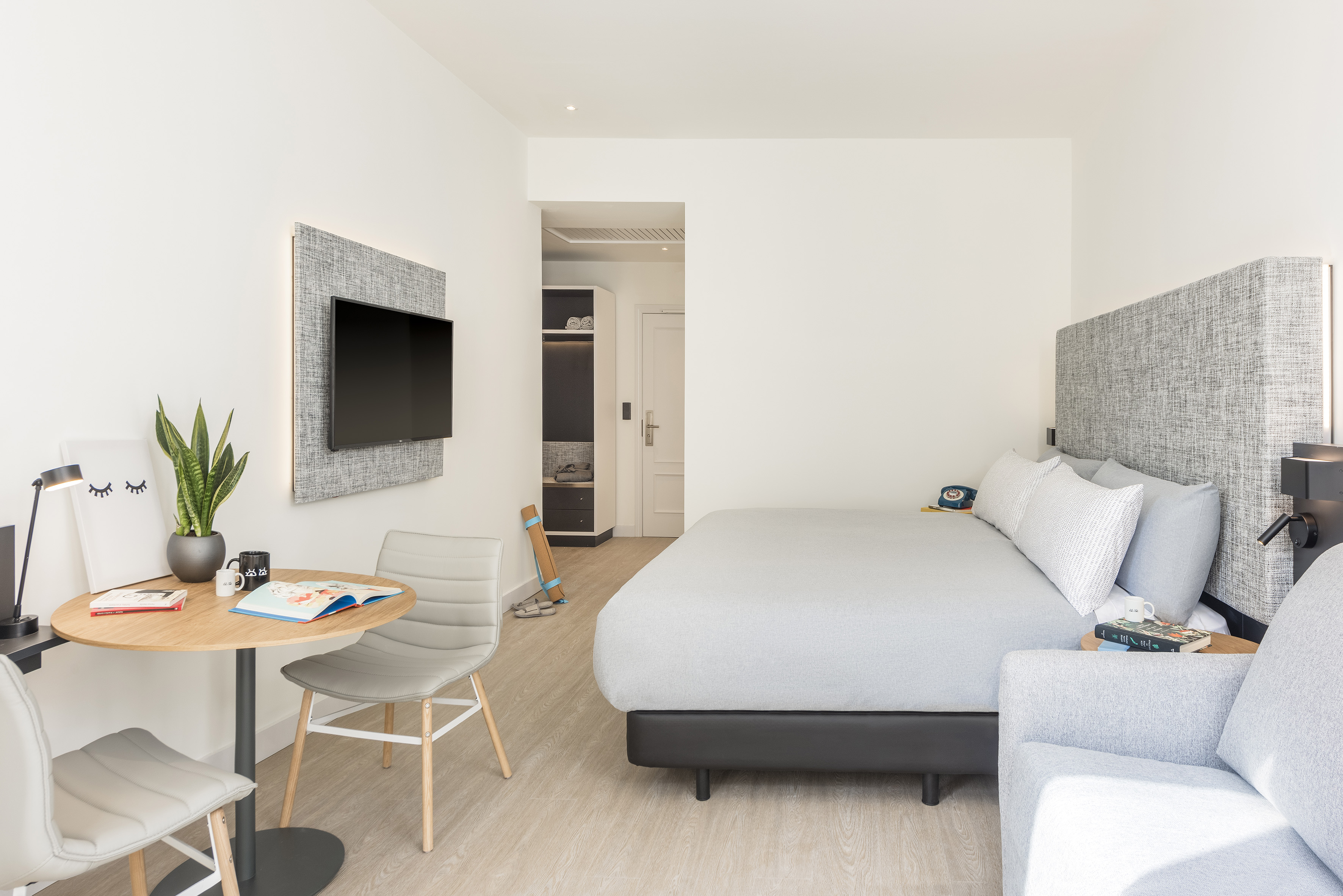 Modern hotel room with gray bed, mounted TV, and small desk area.