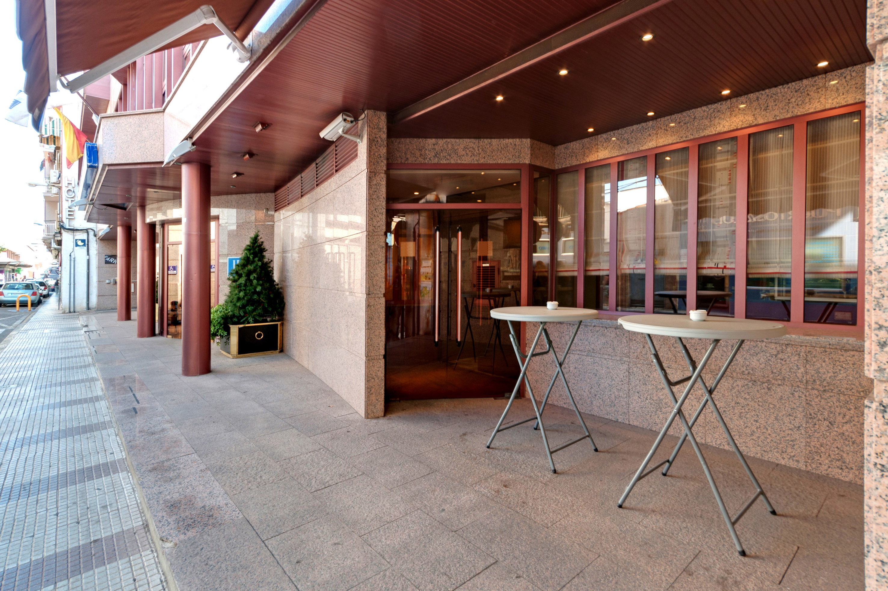 Hotel entrance with two sidewalk tables, red canopy, and large glass windows.