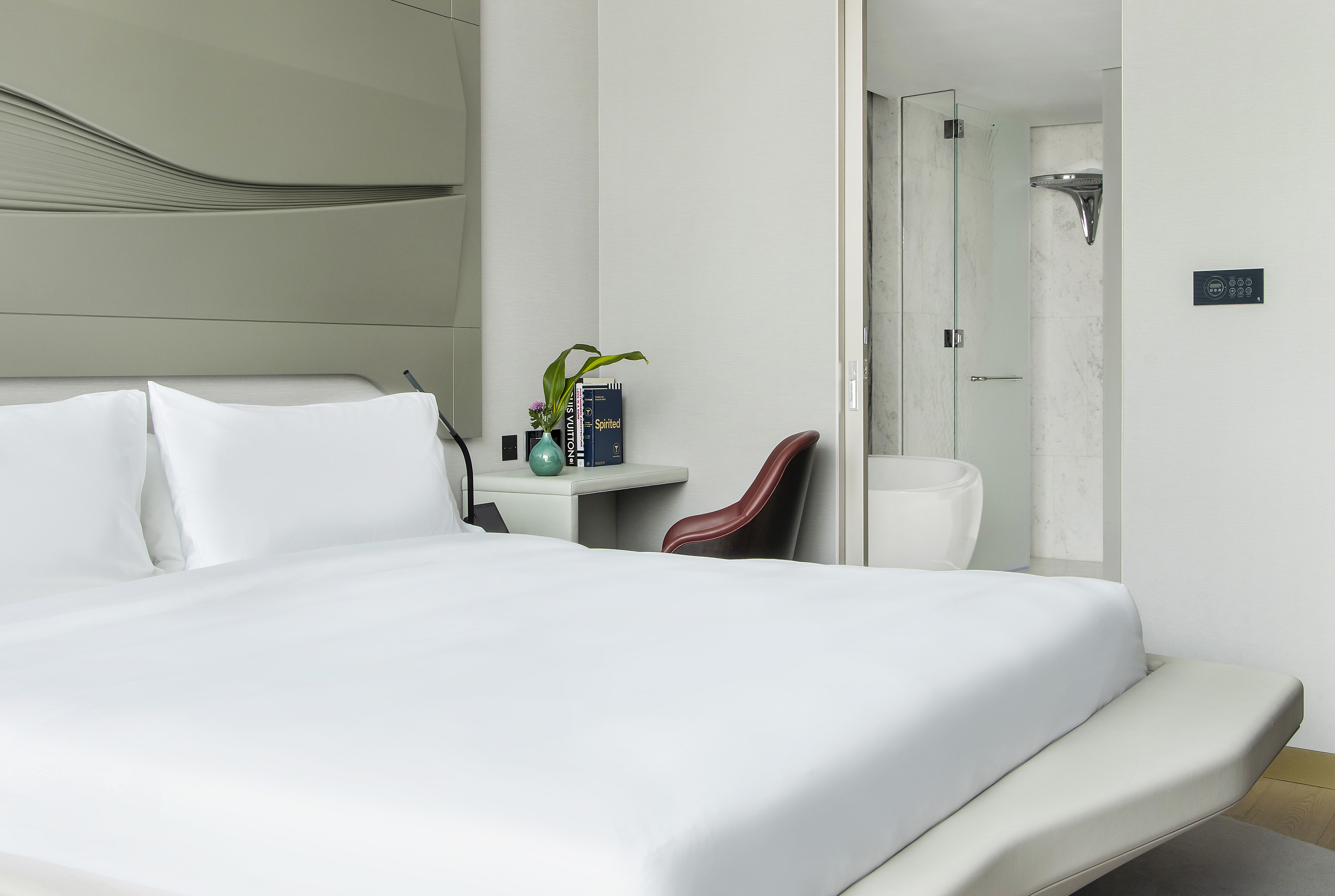 Modern hotel bedroom with white bed, stylish headboard, and glass shower.