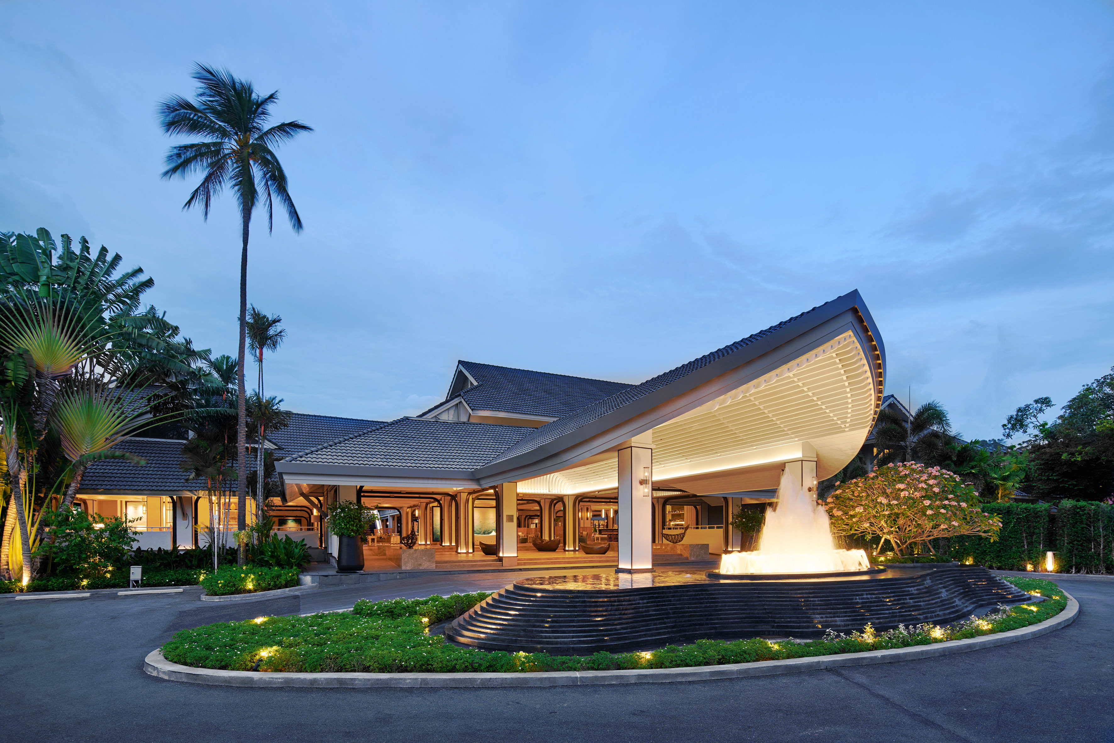 Modern luxury hotel entrance at dusk with illuminated fountain and palm trees.