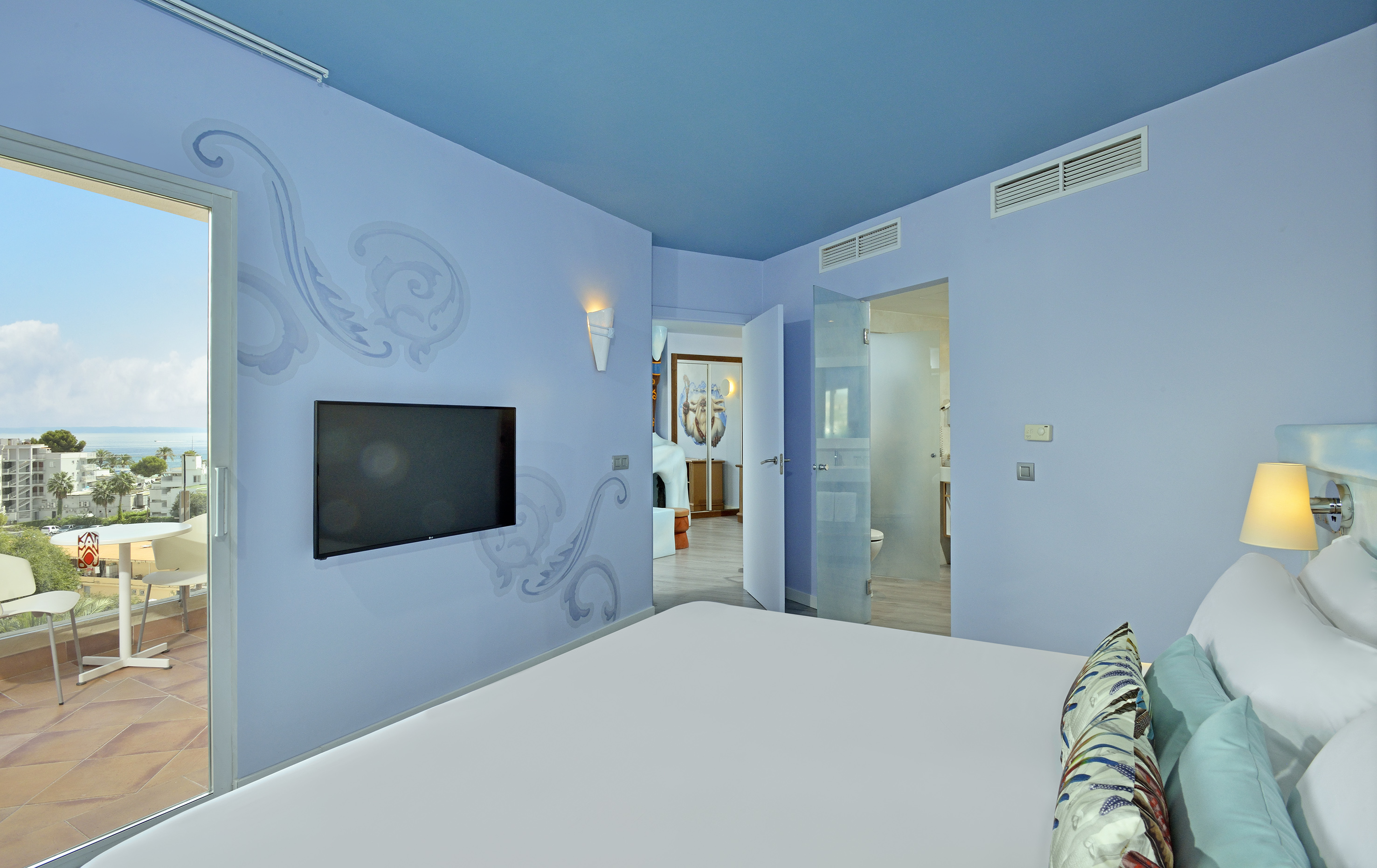 Modern blue hotel bedroom with sea view balcony, TV, and en-suite bathroom.