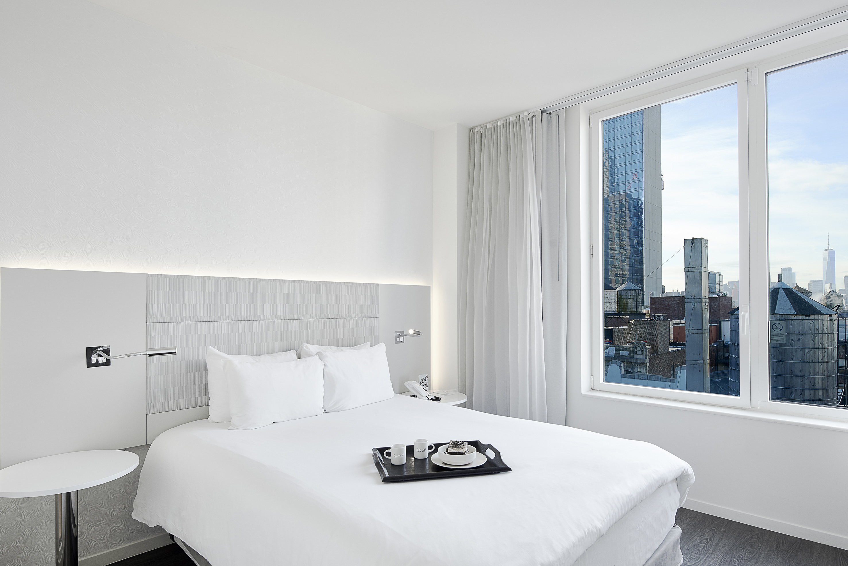 Modern white hotel room, NYC city view, breakfast tray on bed.