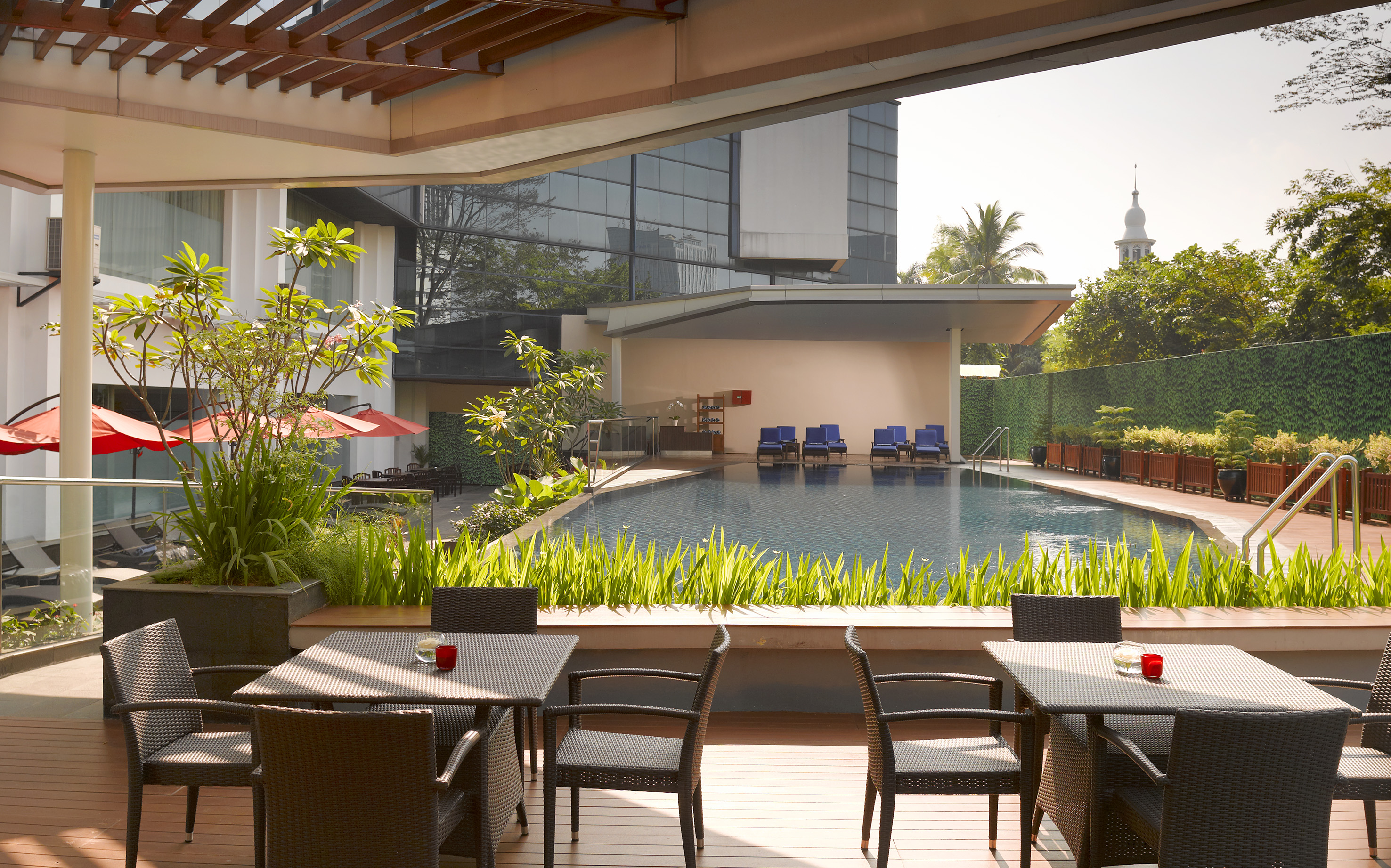 Outdoor hotel pool patio with dining tables, lush greenery, and modern building.