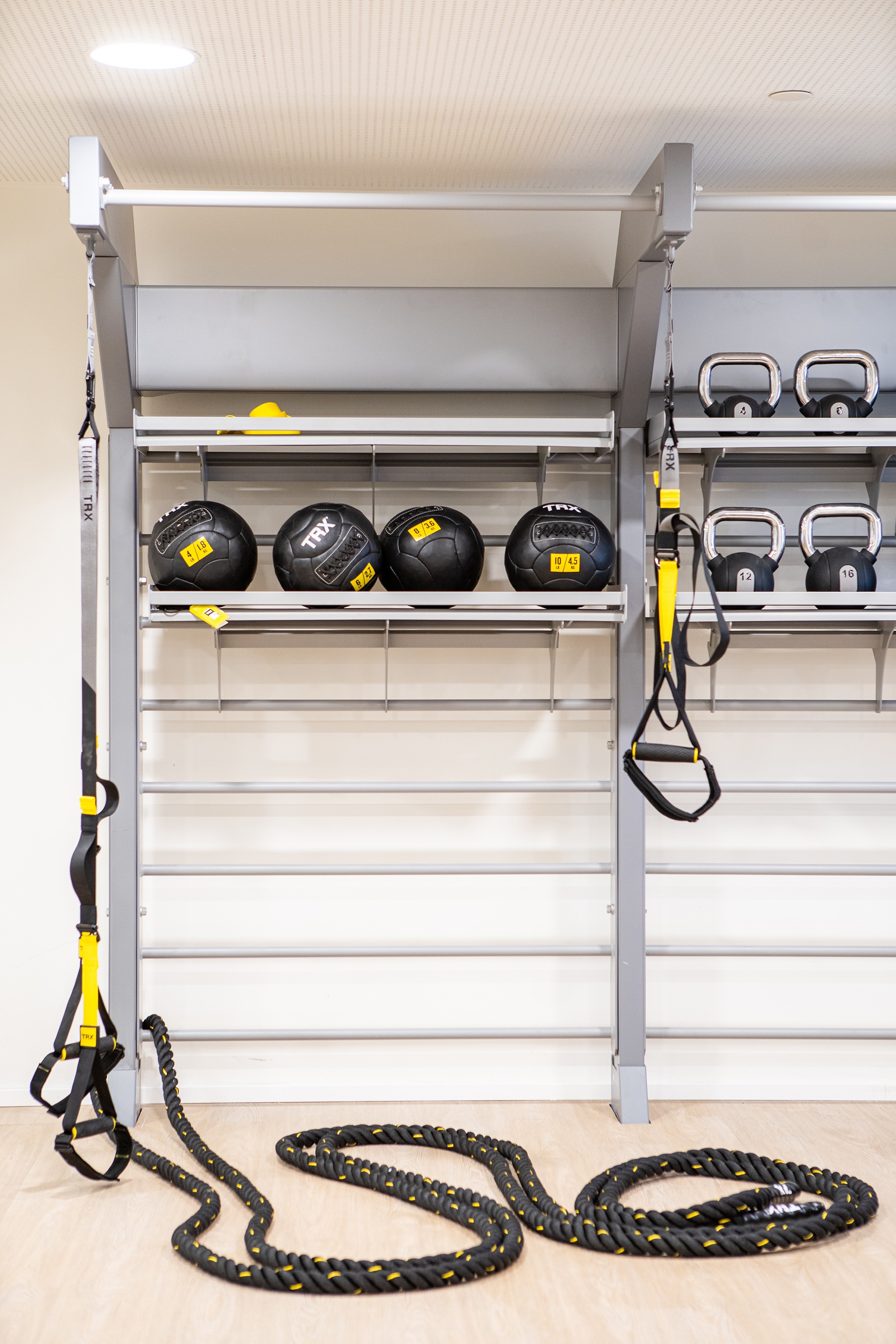 Gym wall rack with medicine balls, kettlebells, TRX trainers, and battle rope.