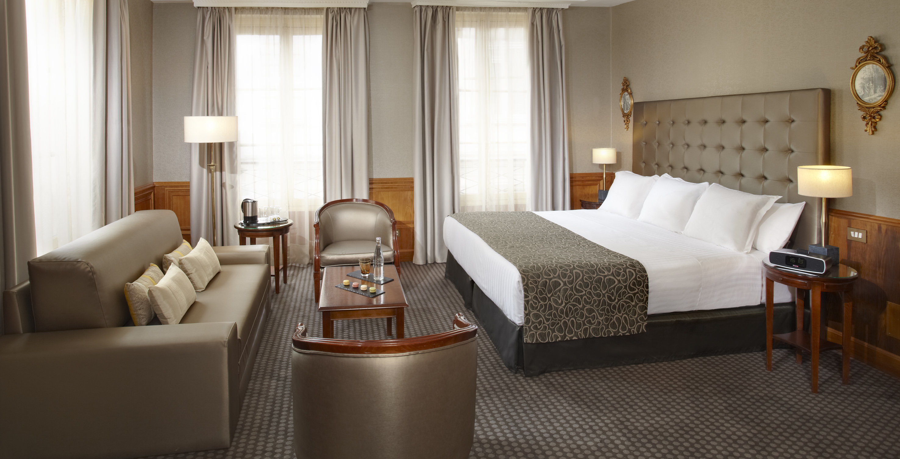 Luxury hotel suite with large bed, sofa, armchairs, and windows.