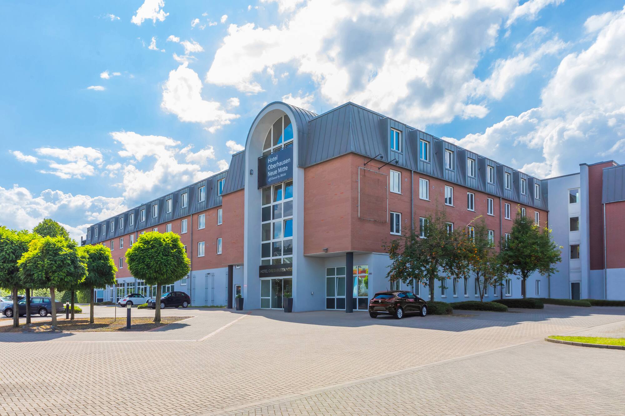 Hotel Oberhausen Neue Mitte exterior with cars parked and trees under a blue sky.