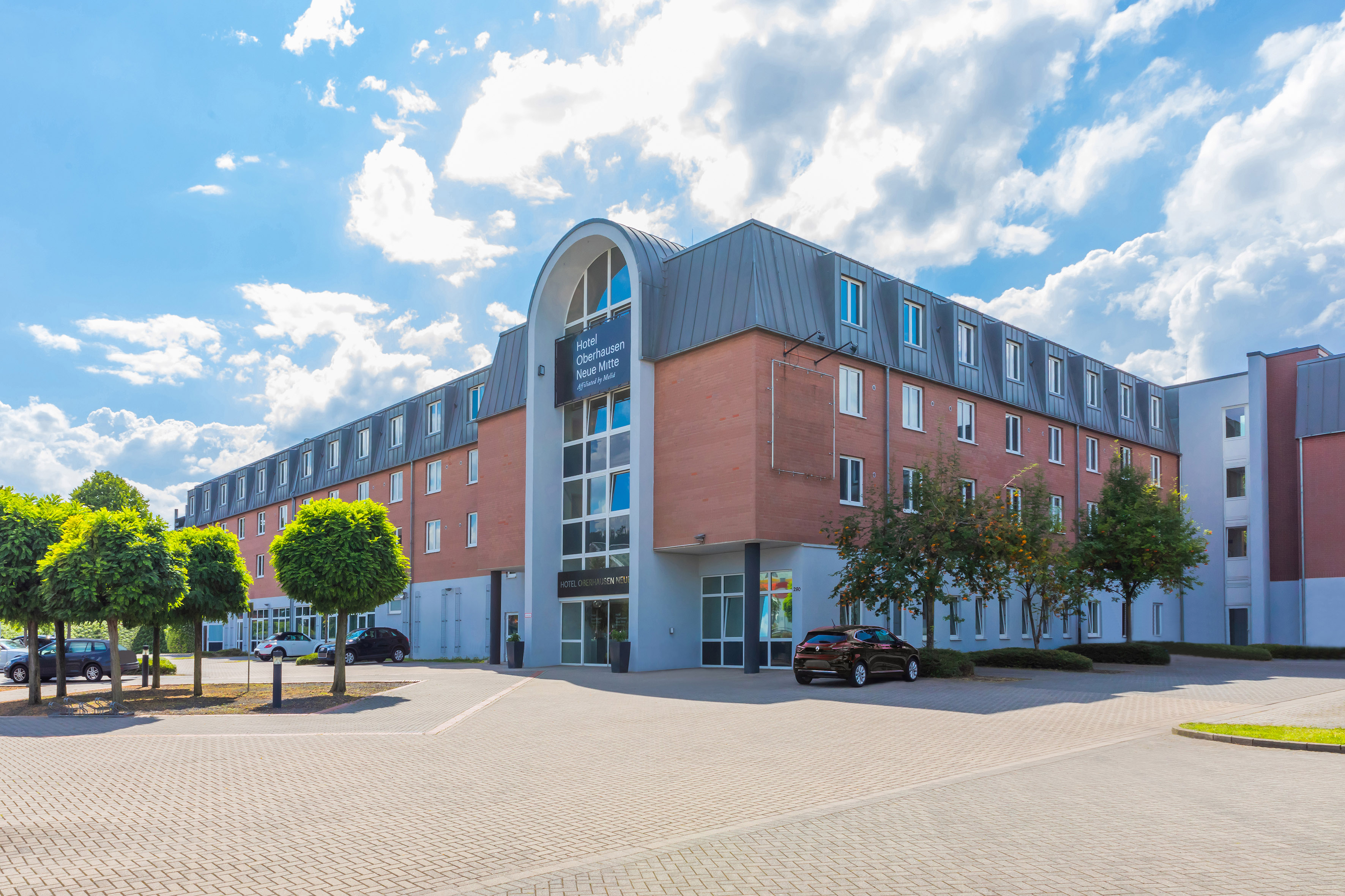 Hotel Oberhausen Neue Mitte exterior with cars parked and trees under a blue sky.