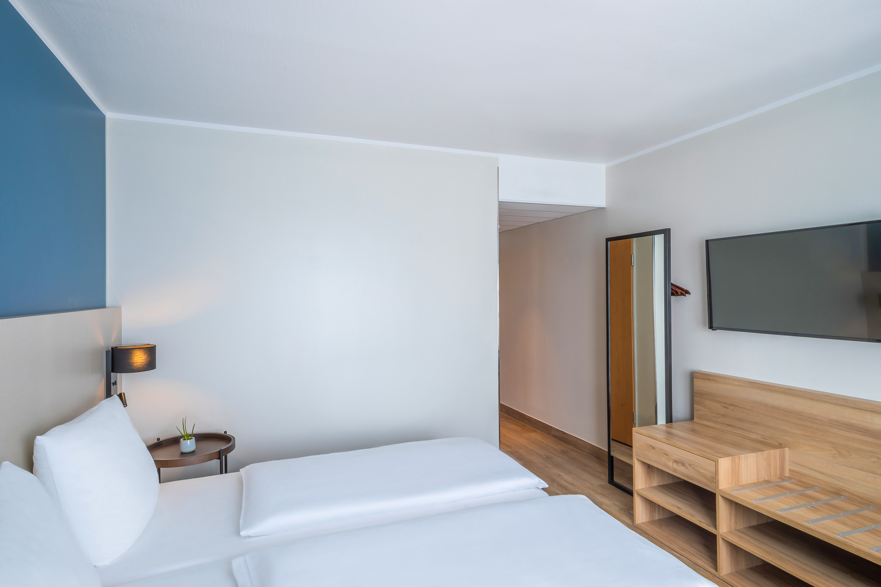Modern hotel room with twin beds, blue accent wall, nightstand, and mounted TV.