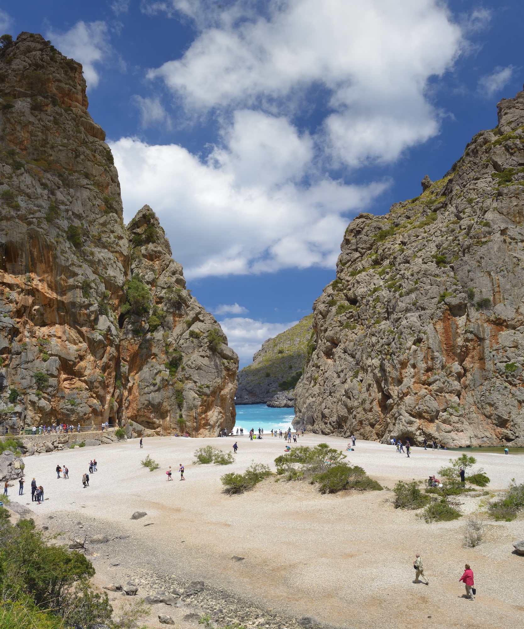 a group of people walking on a sandy beach with large rocks with Samariá Gorge in the background