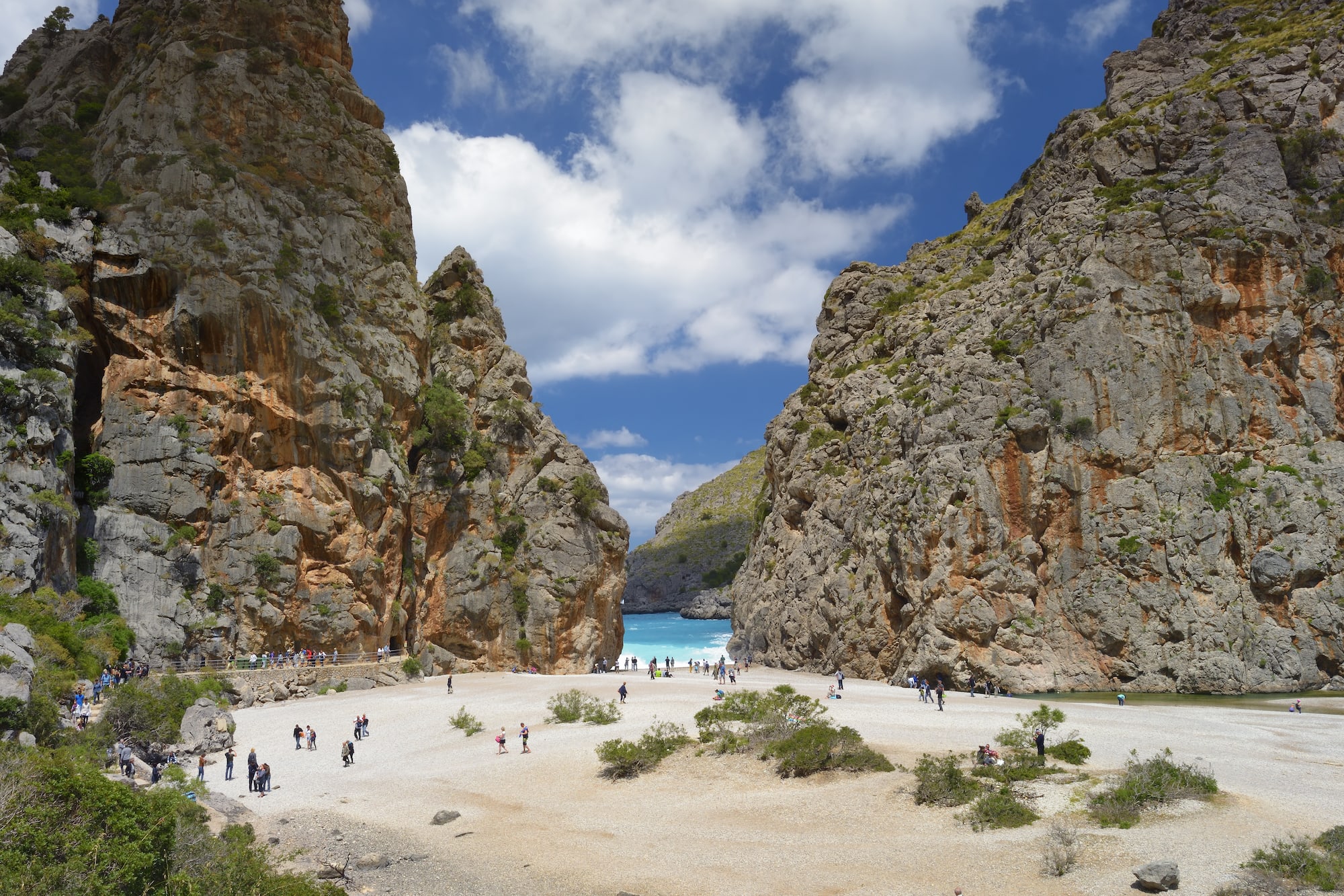 a group of people walking on a sandy beach with large rocks with Samariá Gorge in the background