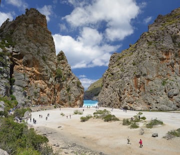 a group of people walking on a sandy beach with large rocks with Samariá Gorge in the background