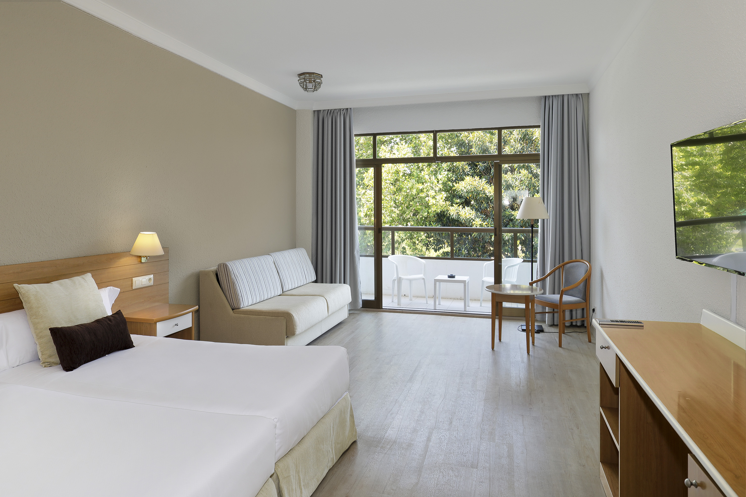 Hotel room with bed, sofa, balcony with outdoor seating, and green nature view.
