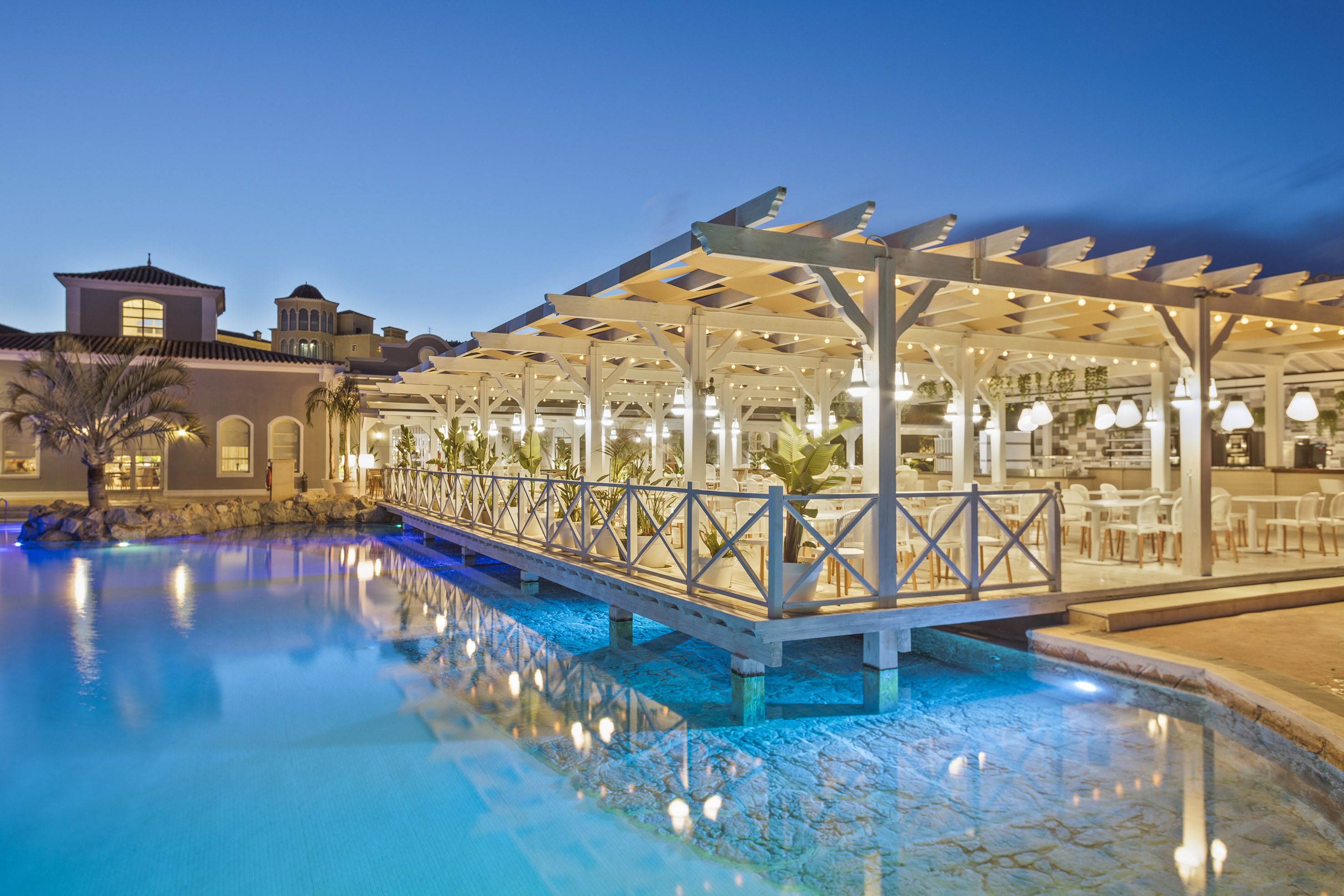 Lit resort restaurant on pier over blue water, white pergola, string lights.