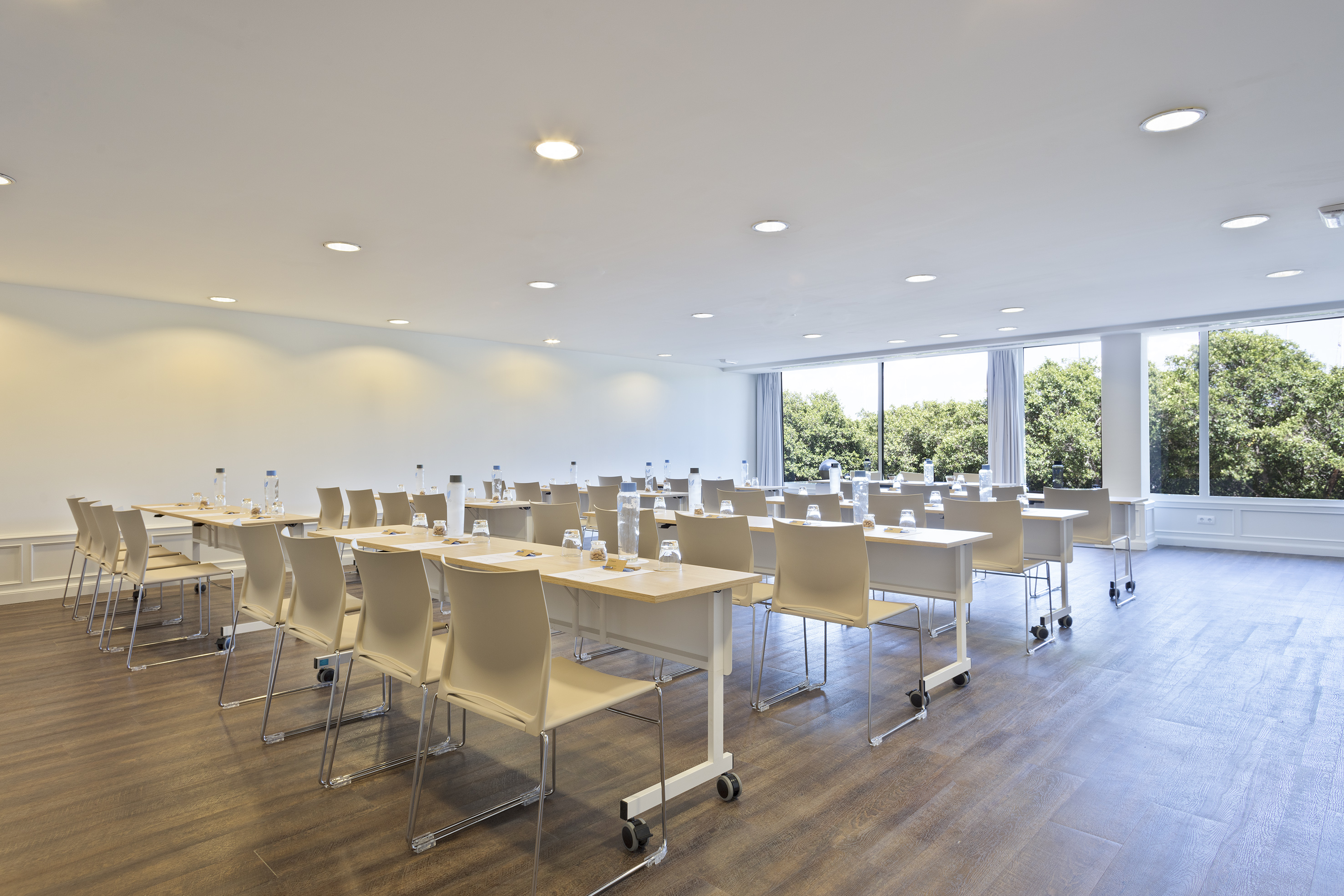 Modern meeting room, light tables and chairs, large windows, green trees.