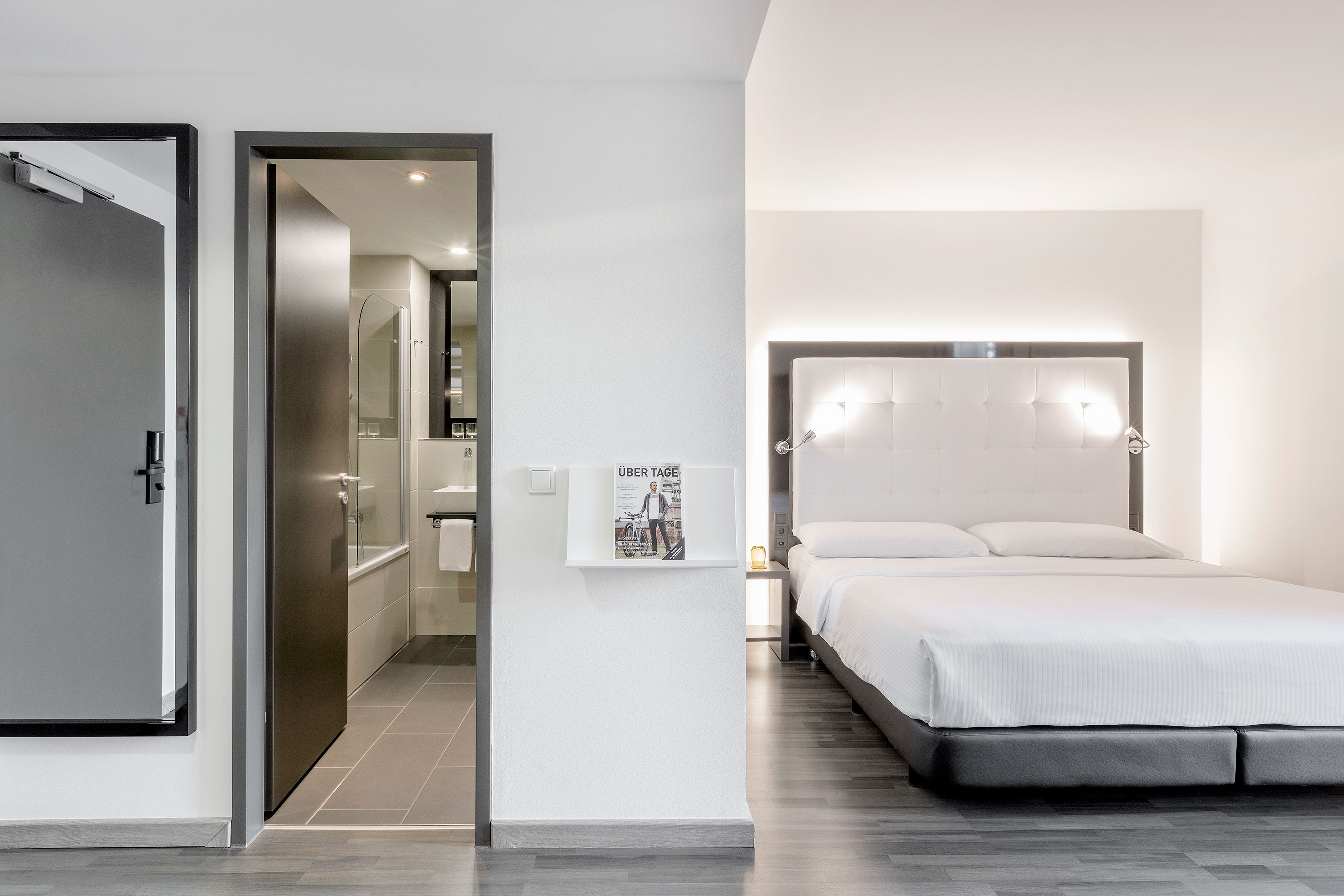 Bright modern hotel bedroom with a white bed, upholstered headboard, and visible bathroom.