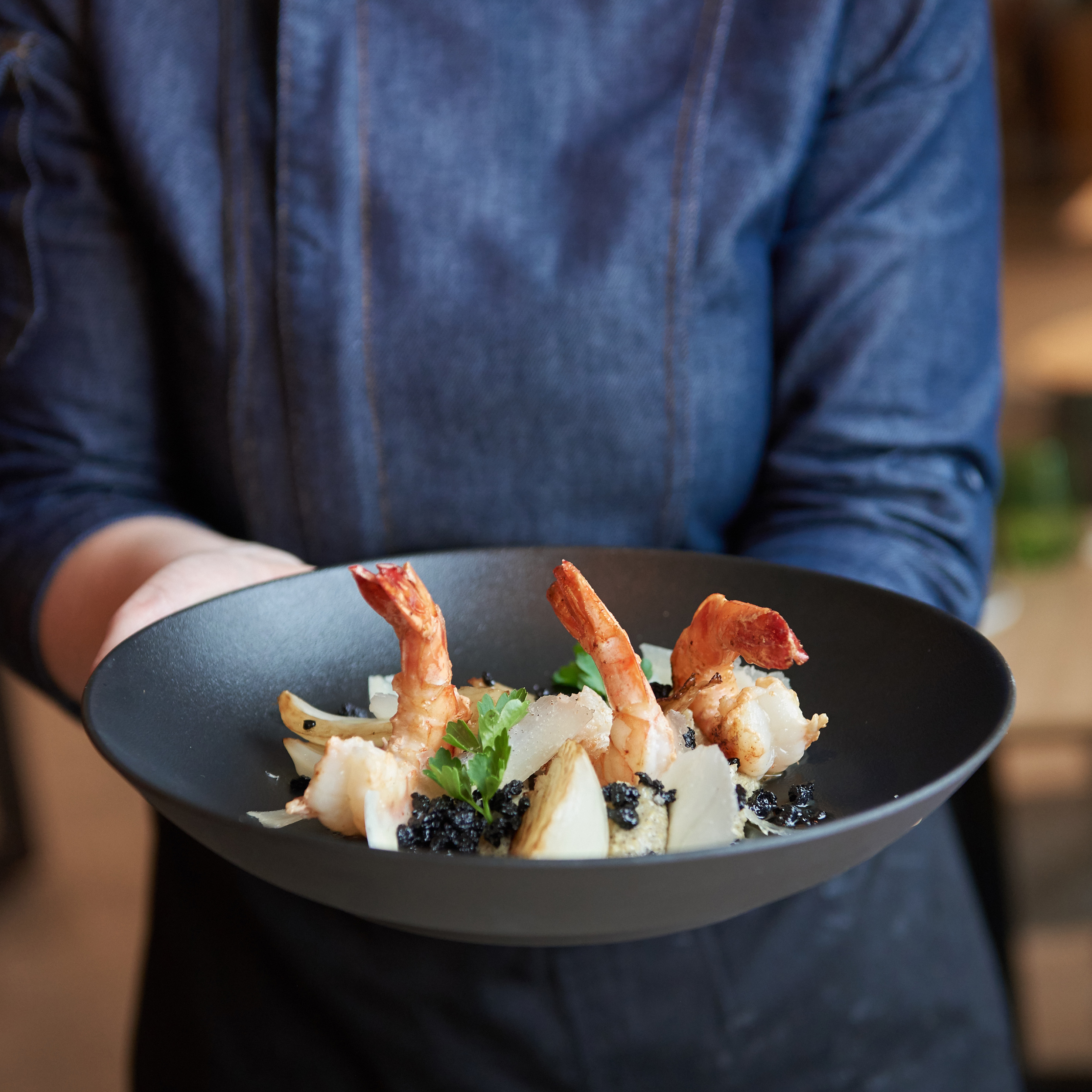Cooked shrimp with caviar and shaved cheese on a black plate, held by a chef.