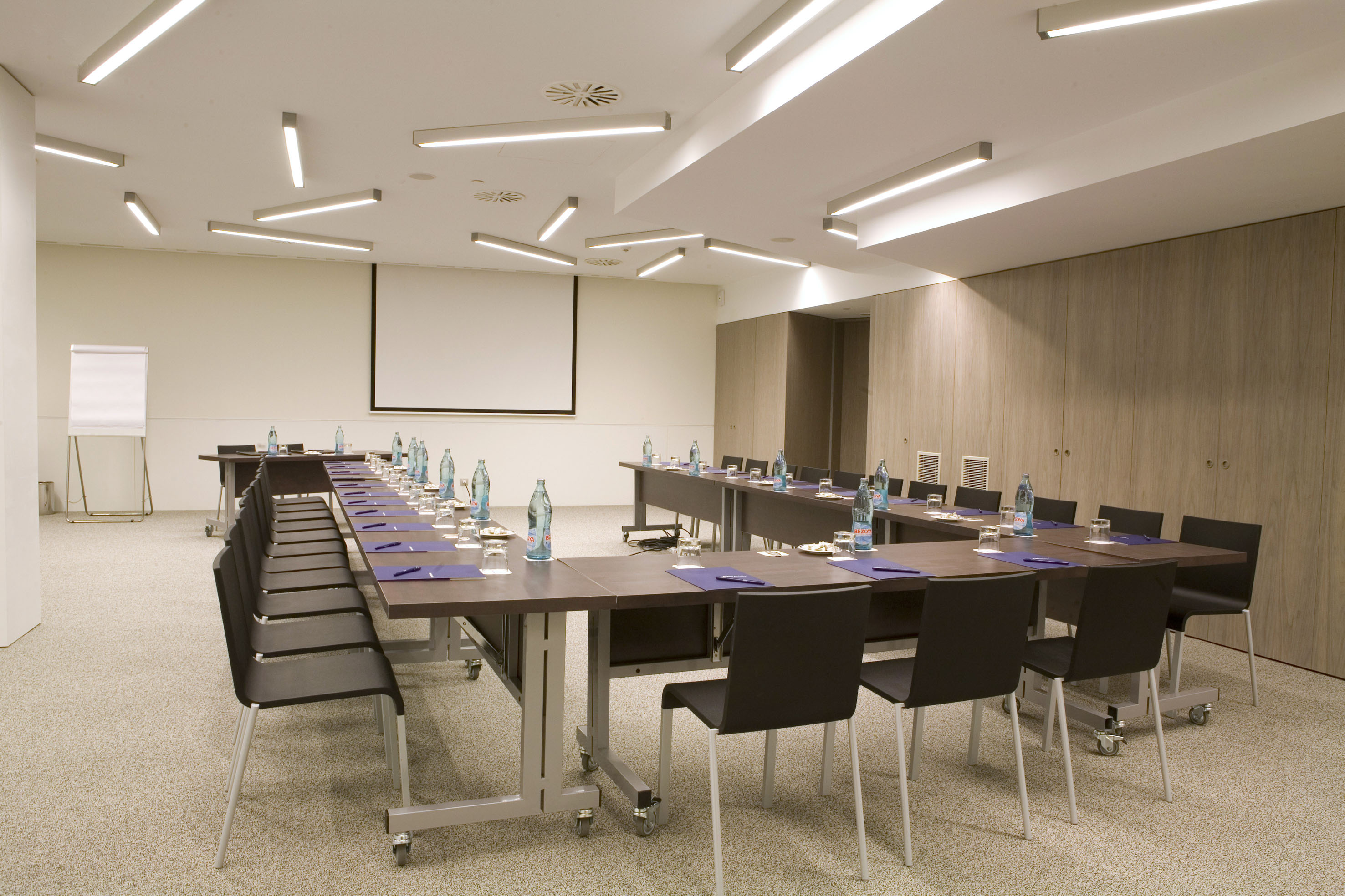 Modern conference room with U-shaped tables, chairs, screen, and whiteboard.