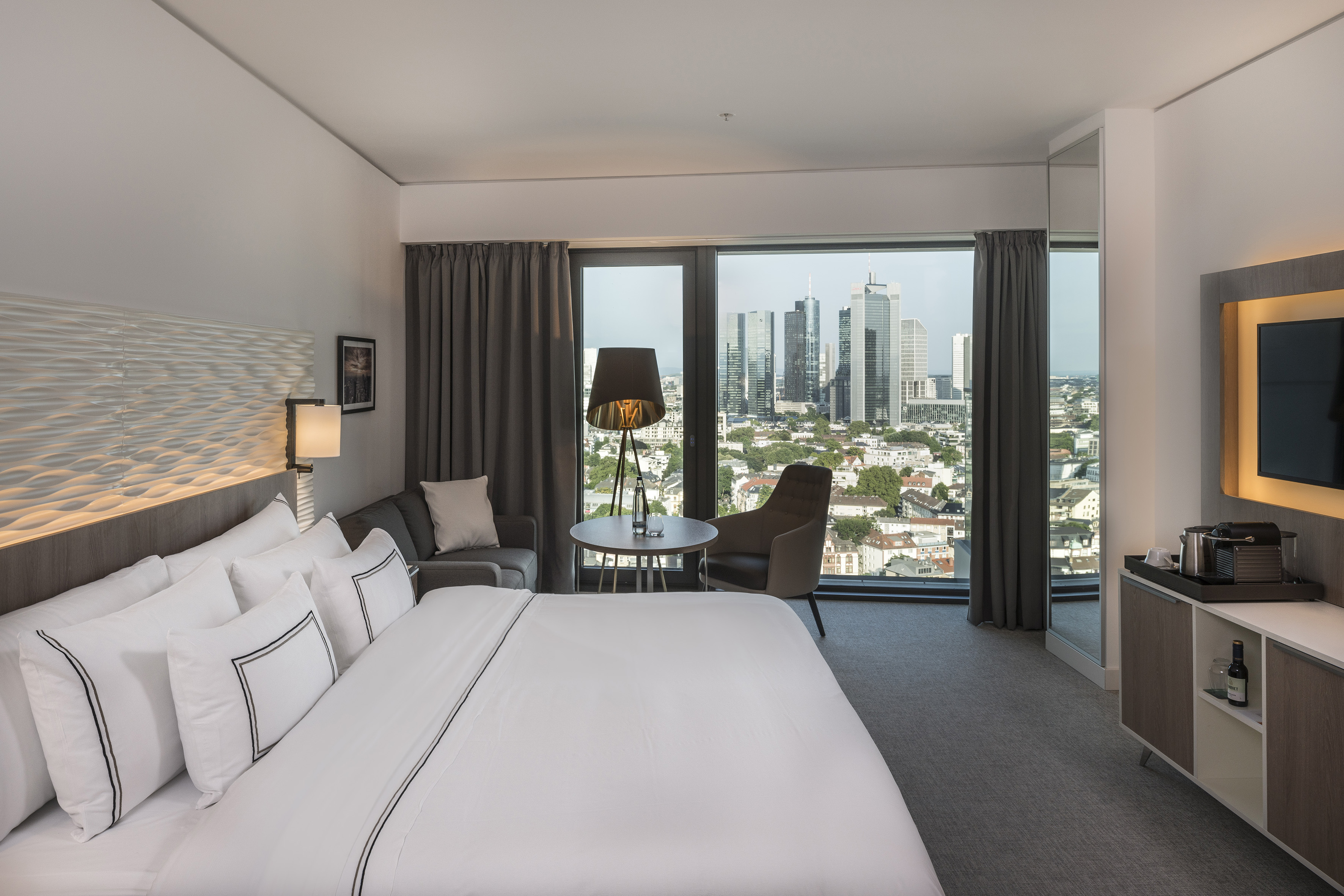 Modern hotel room with king bed, city skyline view, and panoramic windows.