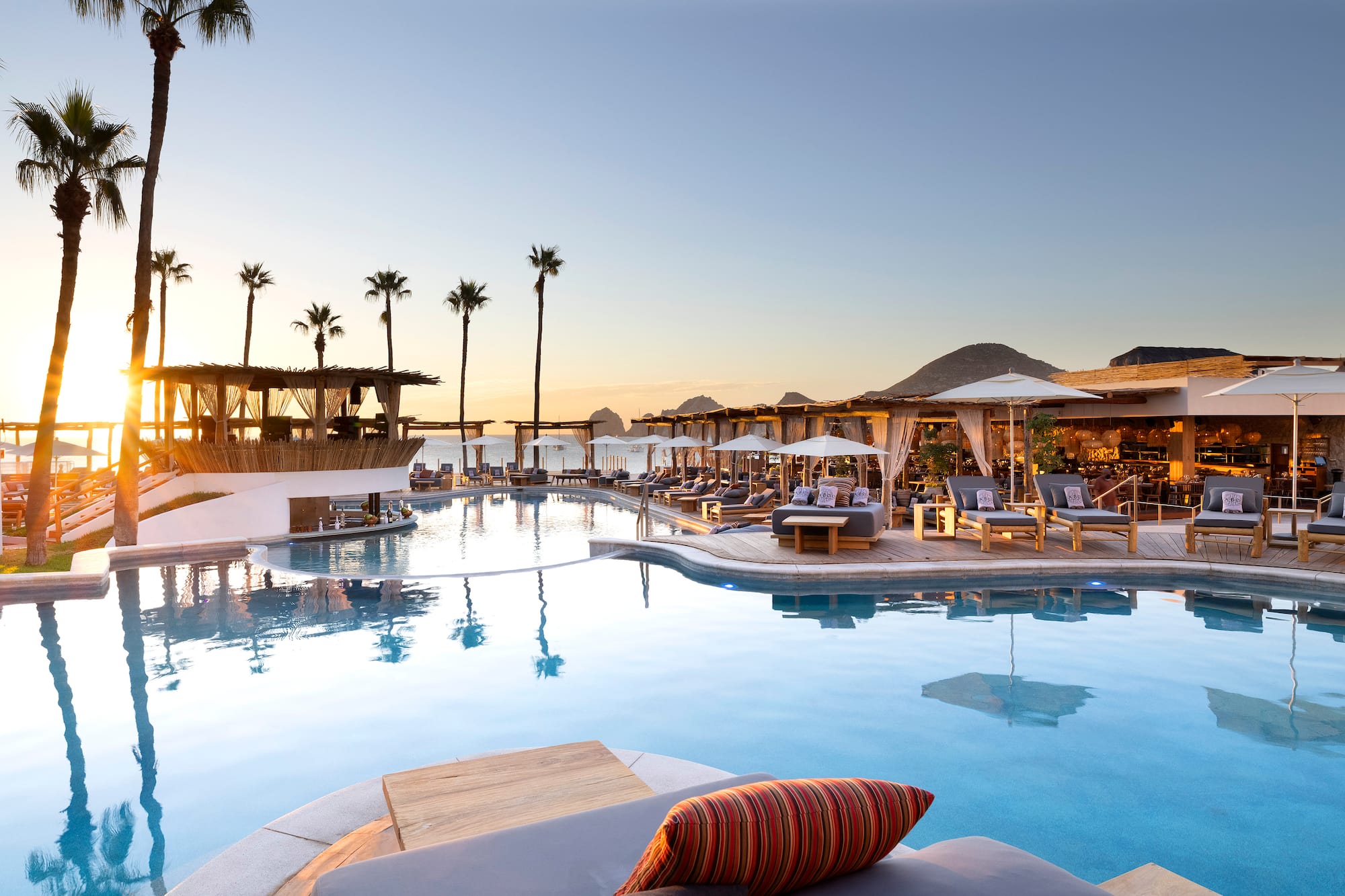 Luxury resort pool at sunset with palm trees, lounge chairs, and cabanas.