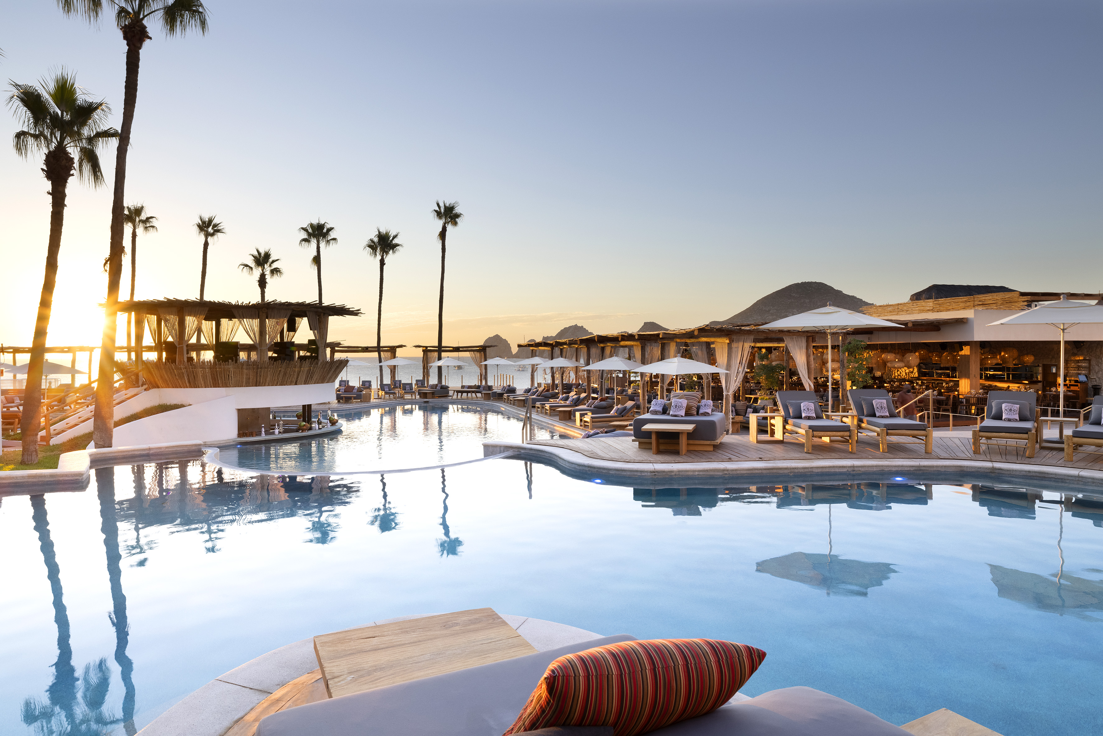 Luxury resort pool at sunset with palm trees, lounge chairs, and cabanas.