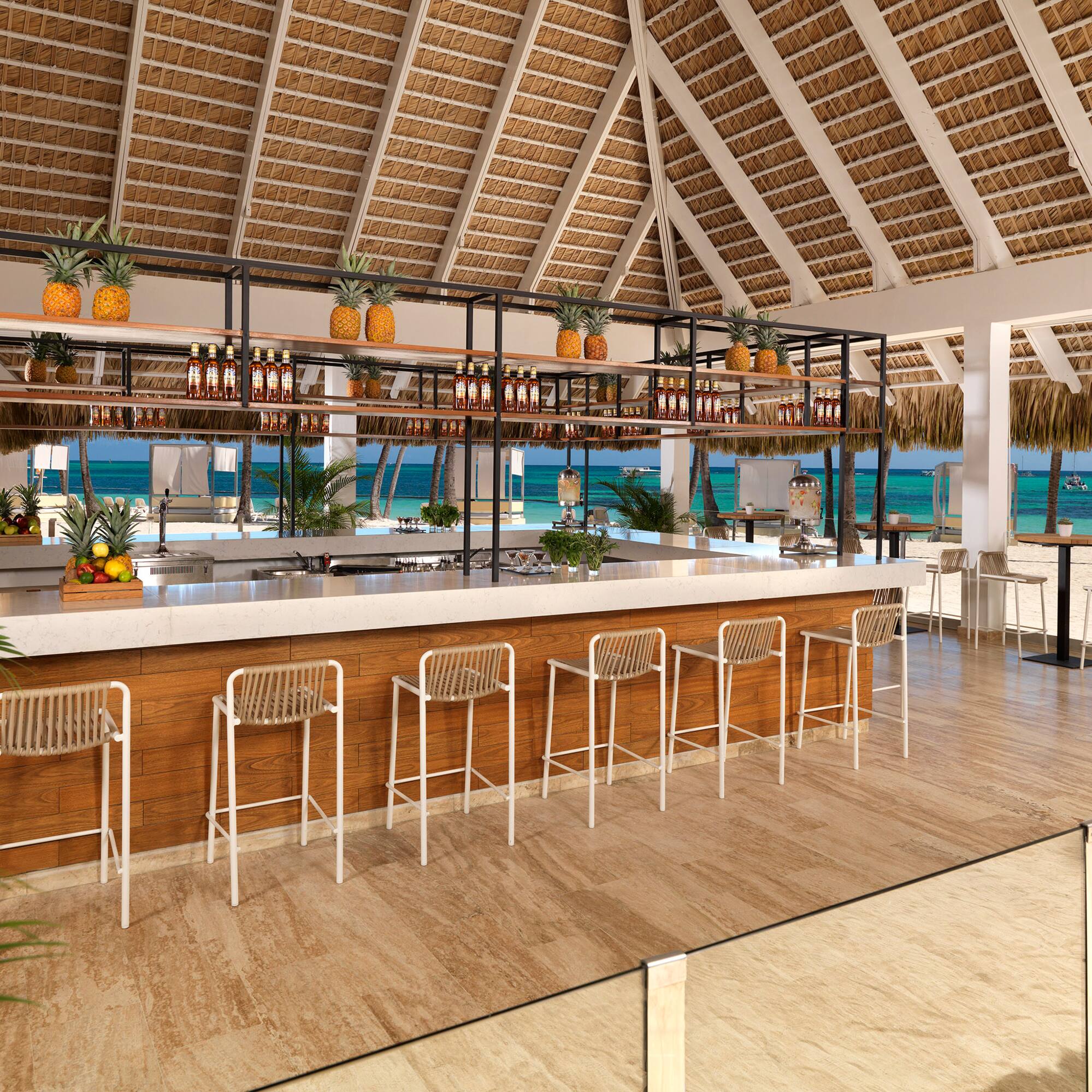 Thatch-roof beach bar, white counter, pineapples, overlooking turquoise ocean.