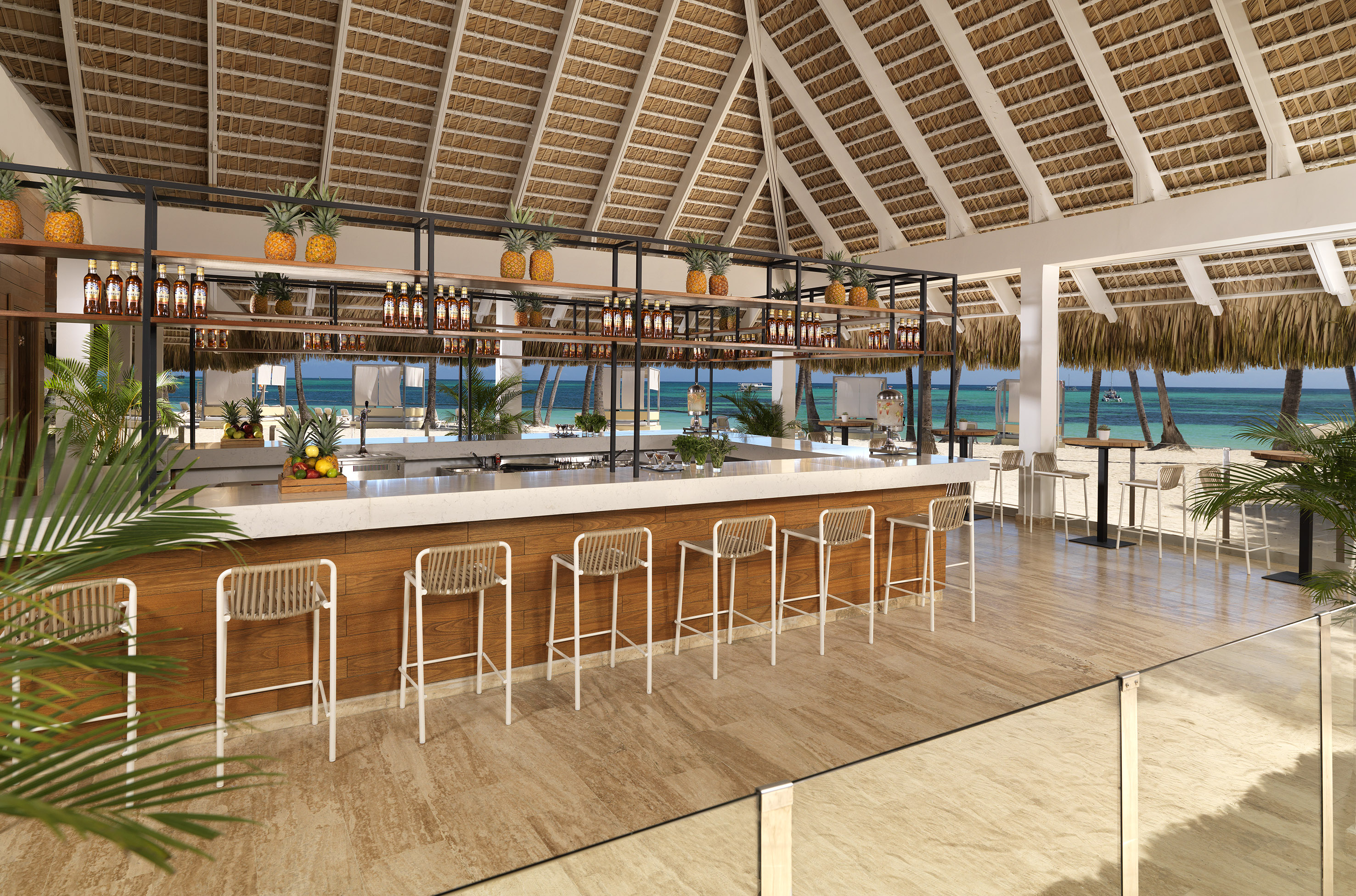 Thatch-roof beach bar, white counter, pineapples, overlooking turquoise ocean.