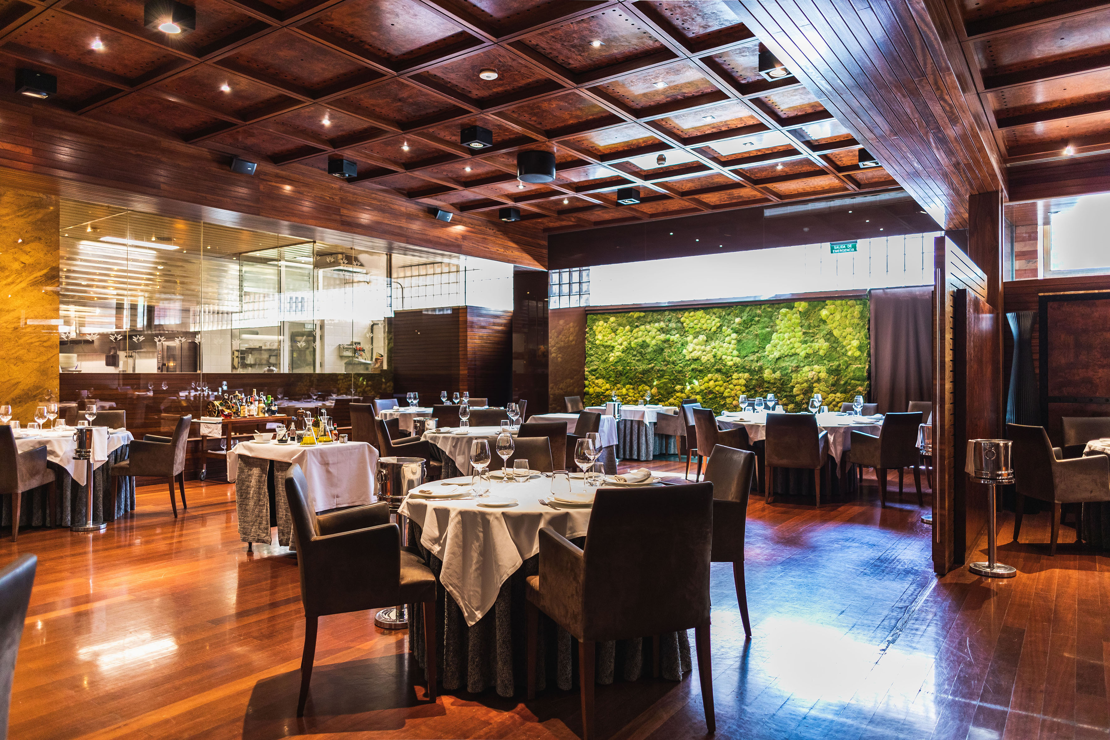 Elegant restaurant dining room with wood decor, moss wall, and open kitchen.