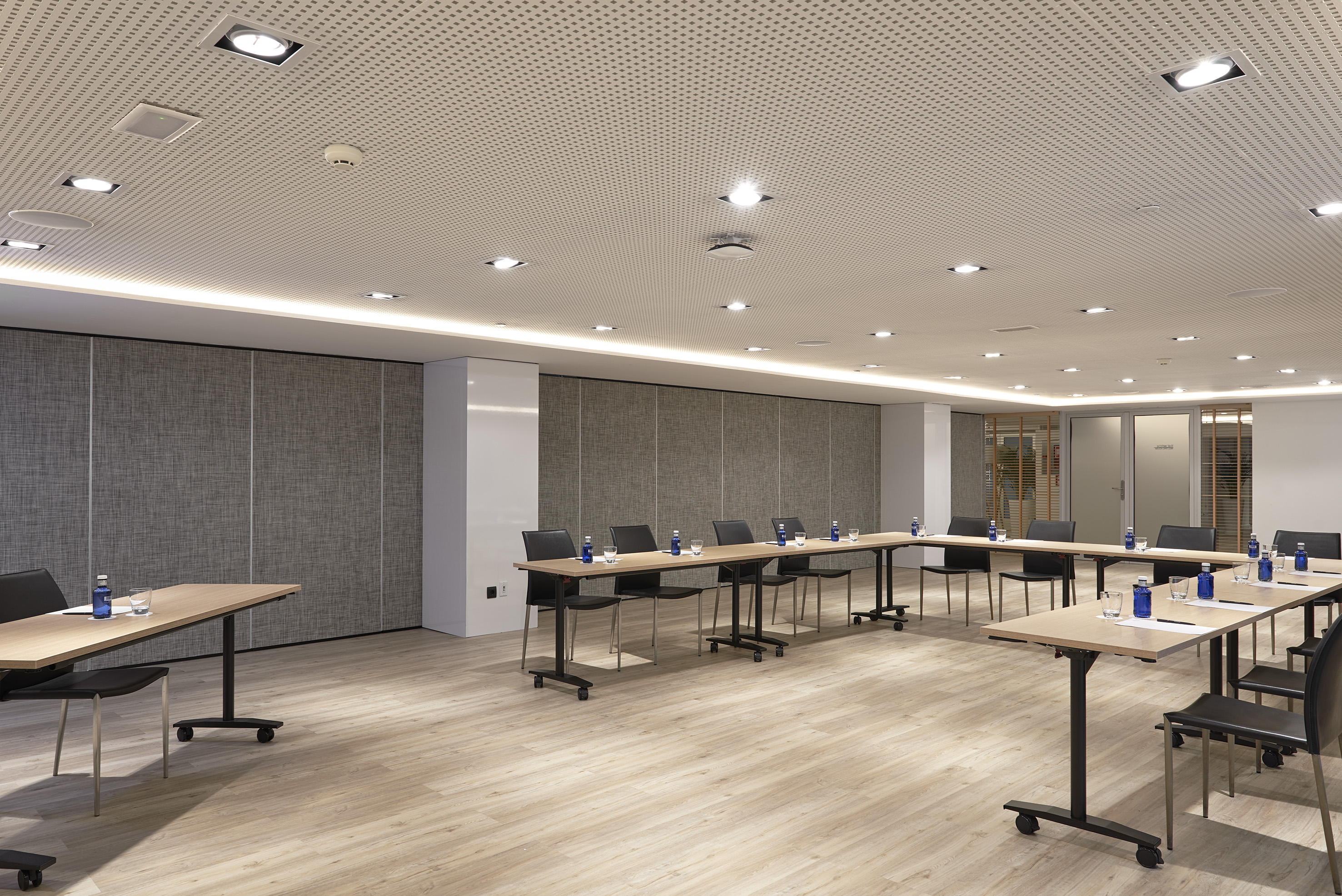 Modern meeting room with U-shaped tables, wood floor, and perforated ceiling.