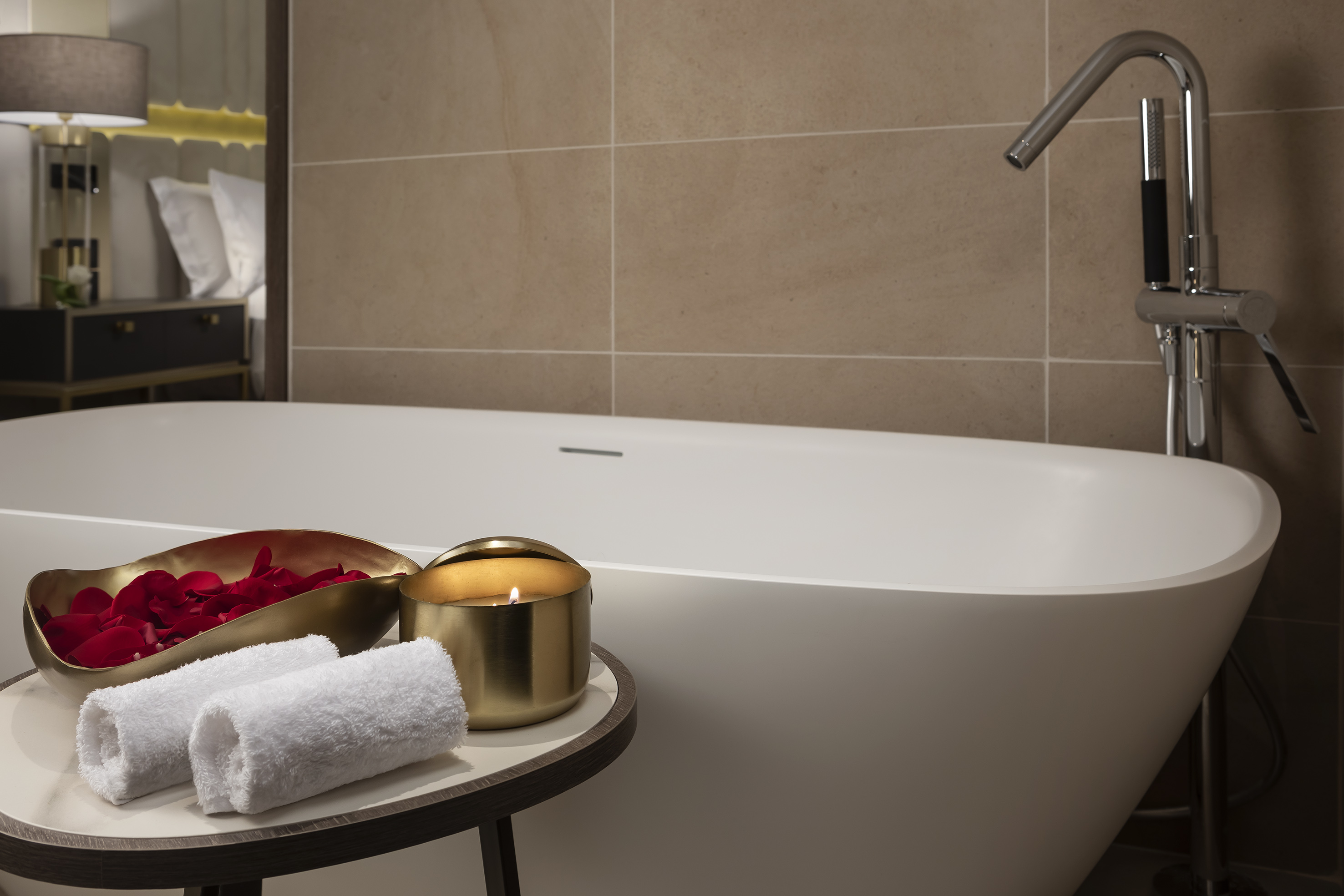 Luxury hotel bath setup with freestanding tub, rose petals, candle, towels.