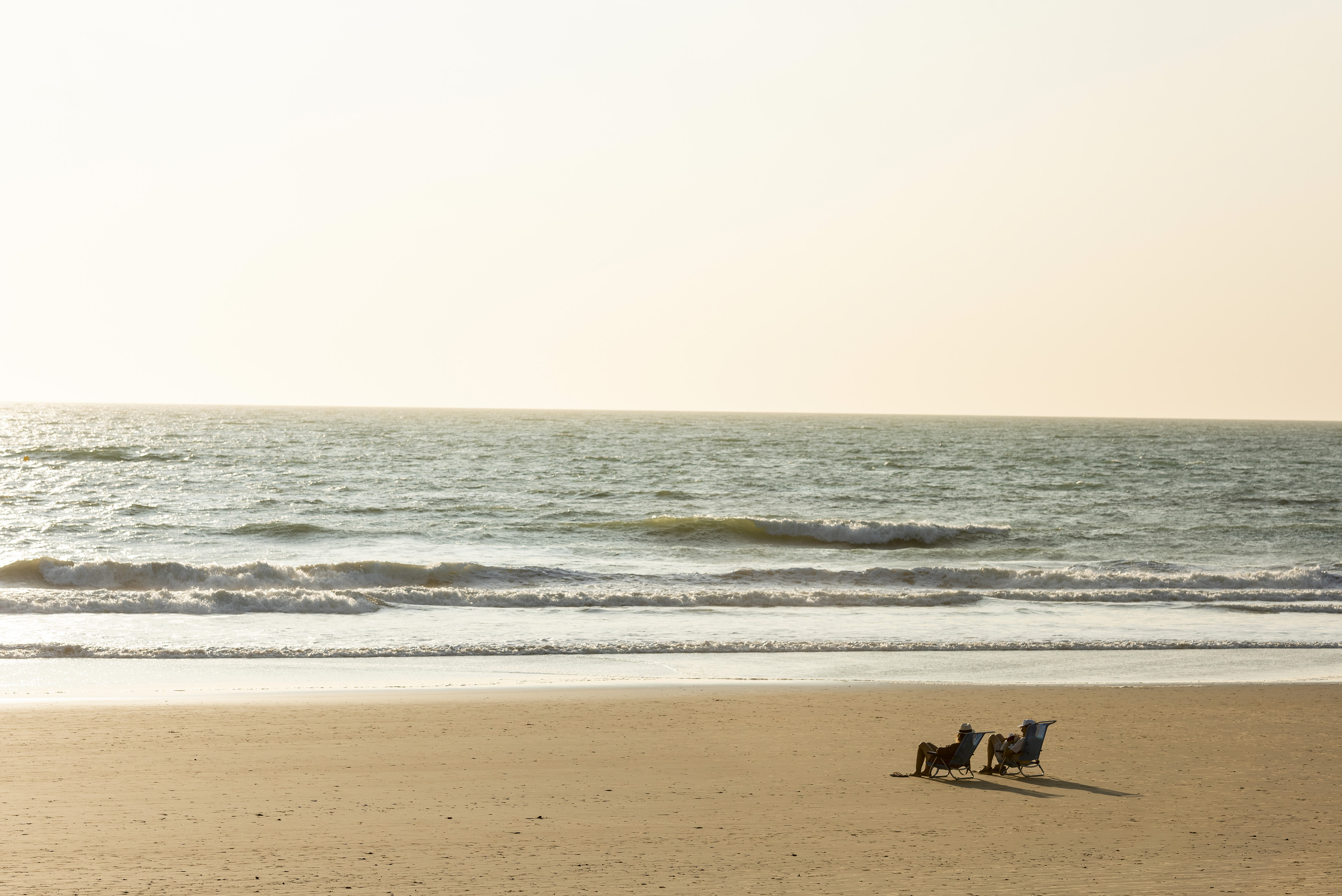 Couple relaxing in beach chairs on sandy shore facing ocean waves.
