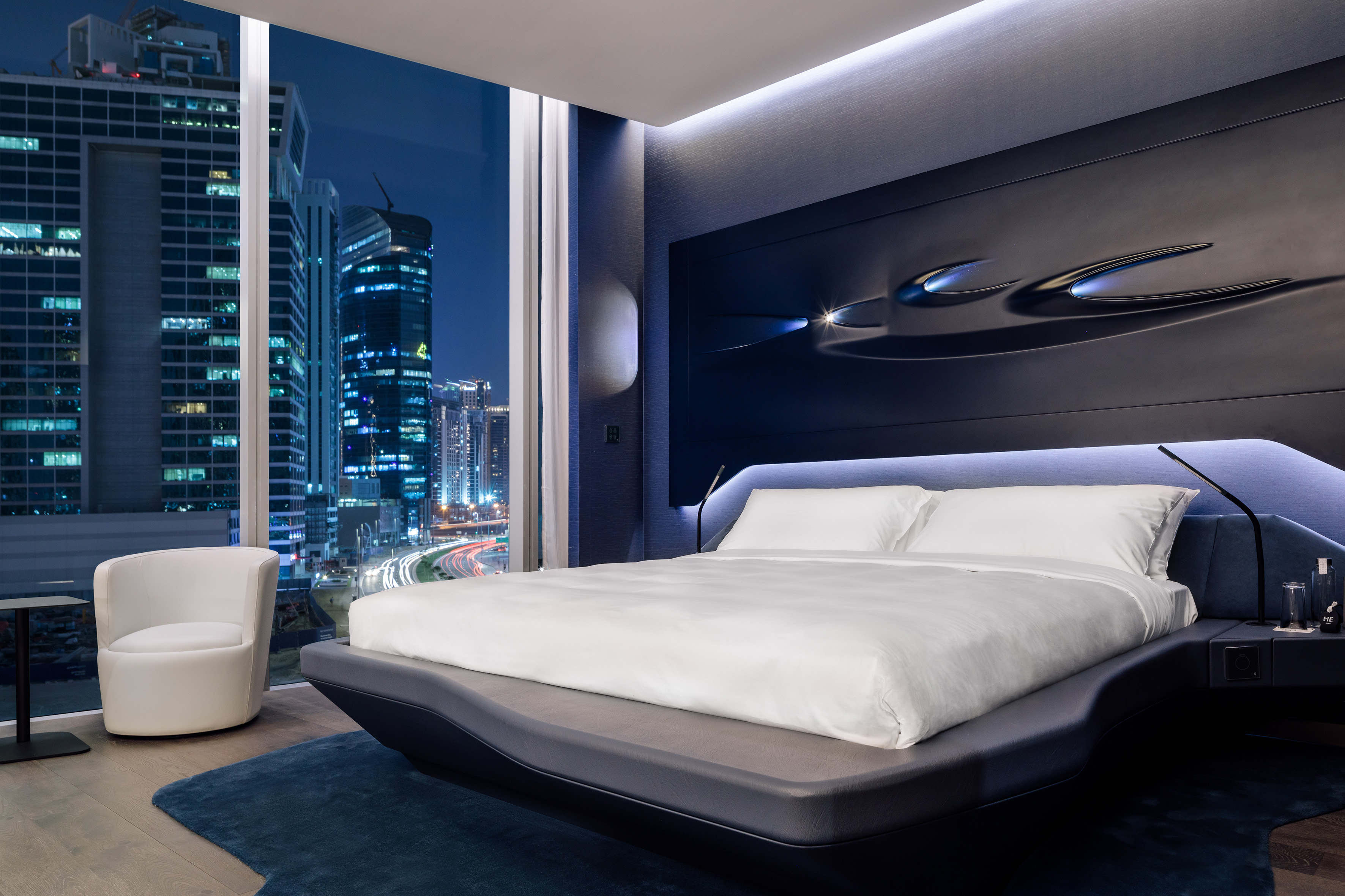 Modern hotel bedroom with city night view, blue design, large window.