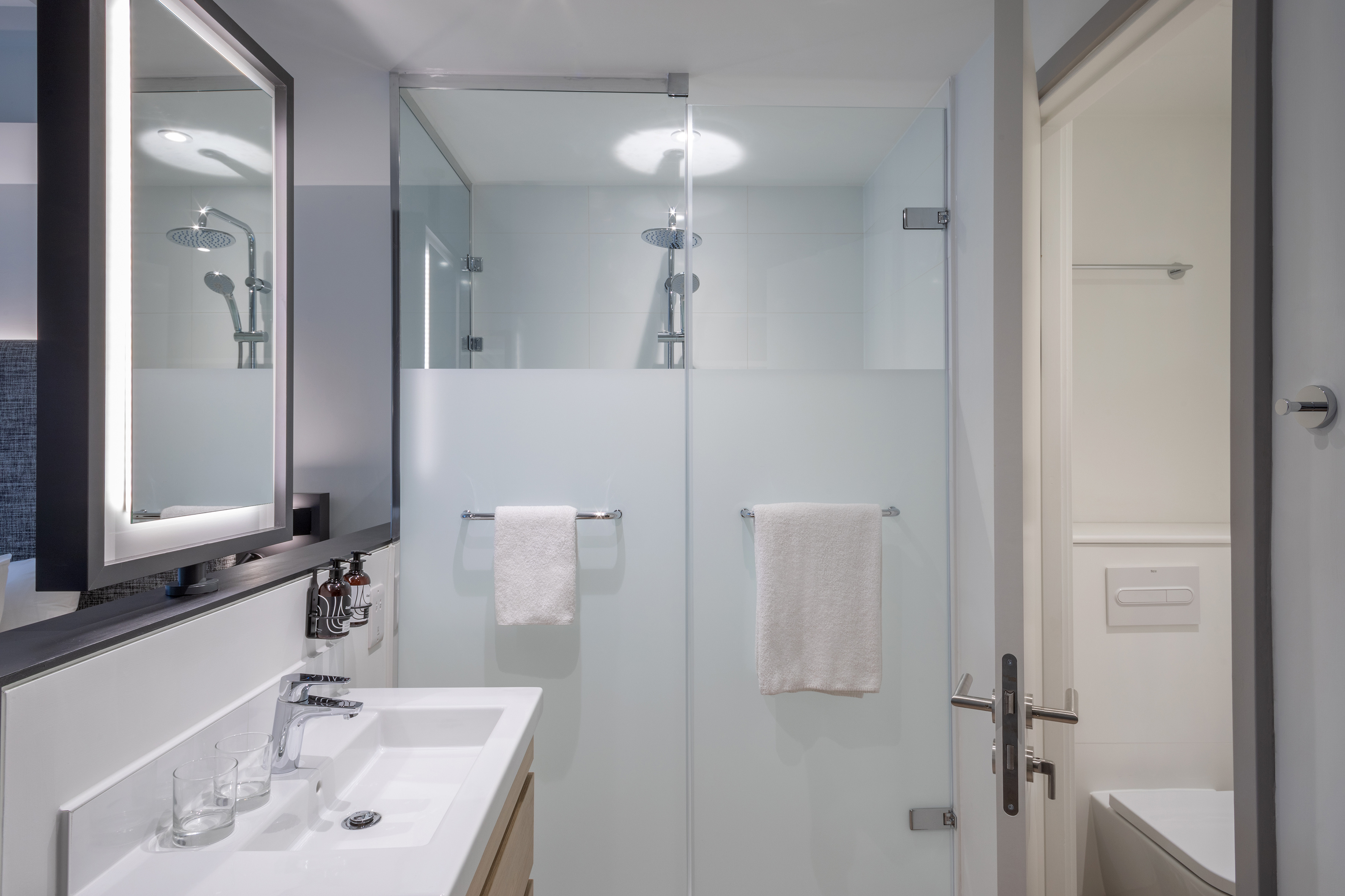 Bright hotel bathroom with modern vanity, backlit mirror, and glass shower.