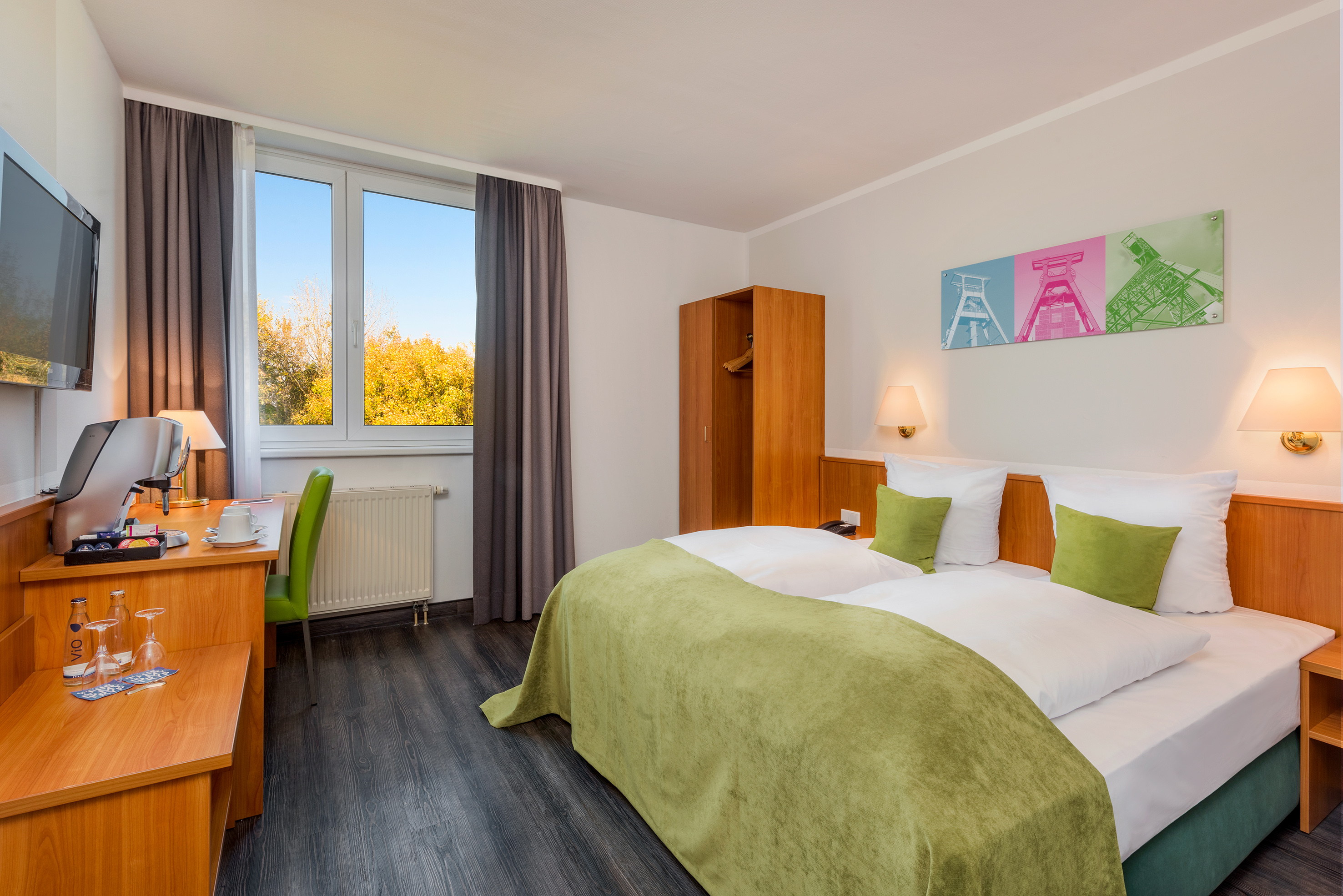 Hotel room with green bed, desk, autumn view, and industrial mining art.
