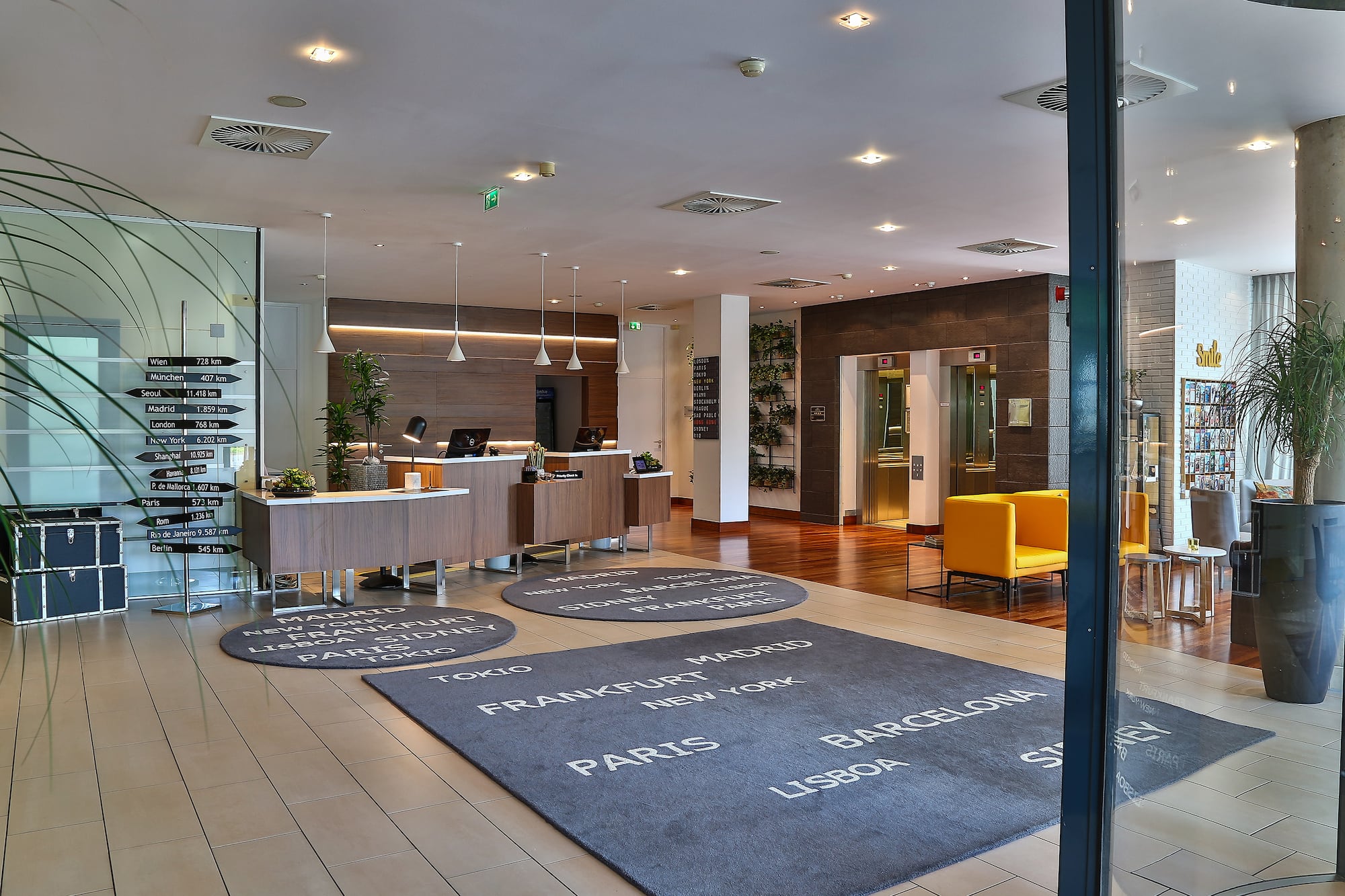Modern hotel lobby with reception, travel destination sign, and world city rugs.
